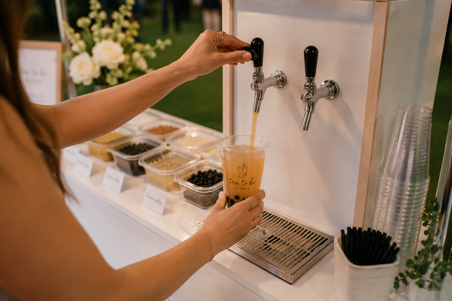 A person is pouring a beverage from a tap into a plastic cup on a white counter at a Tea to Be self serve bubble tea cart, with toppings in small containers nearby.