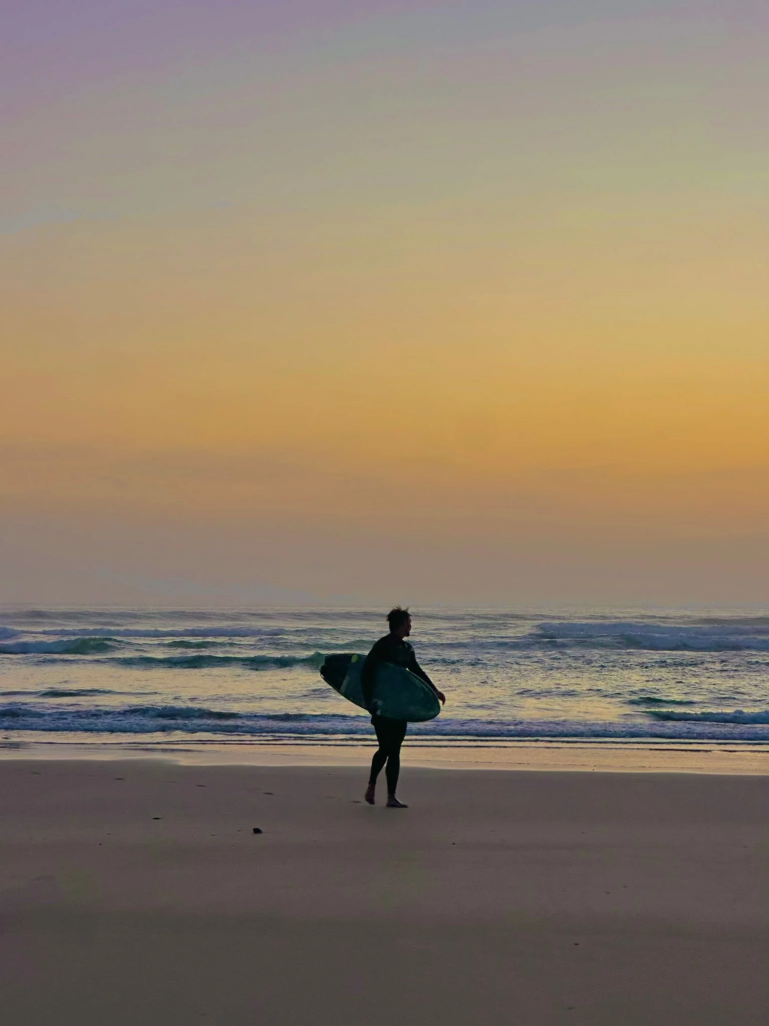 Surfer (Logan) carrying a board along the shore at sunset in Cornwall.