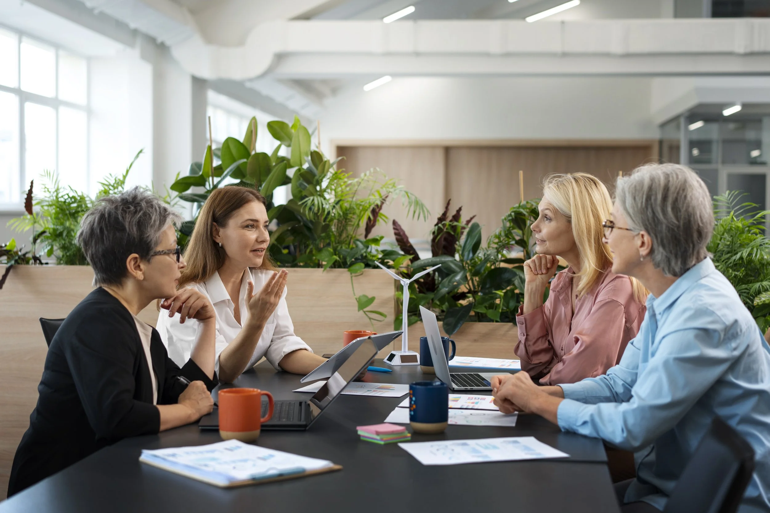 Five women having a discussion in a modern office with lush green plants and large windows.