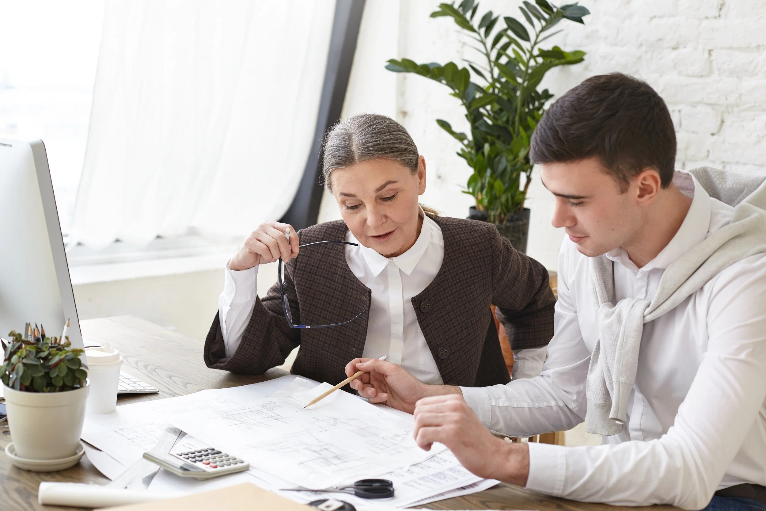 Two people, a woman and a man, working together at a desk covered with papers, a calculator, and drawing tools, in a bright room with a window and a large green plant in the background.