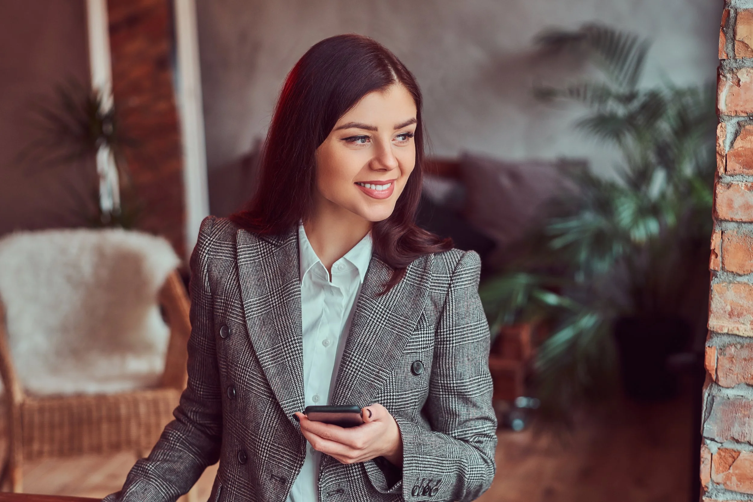 A woman with long, dark red hair and light skin, wearing a gray plaid blazer and white shirt, sitting in a cozy, rustic cafe. She is holding a phone in her right hand and smiling softly.