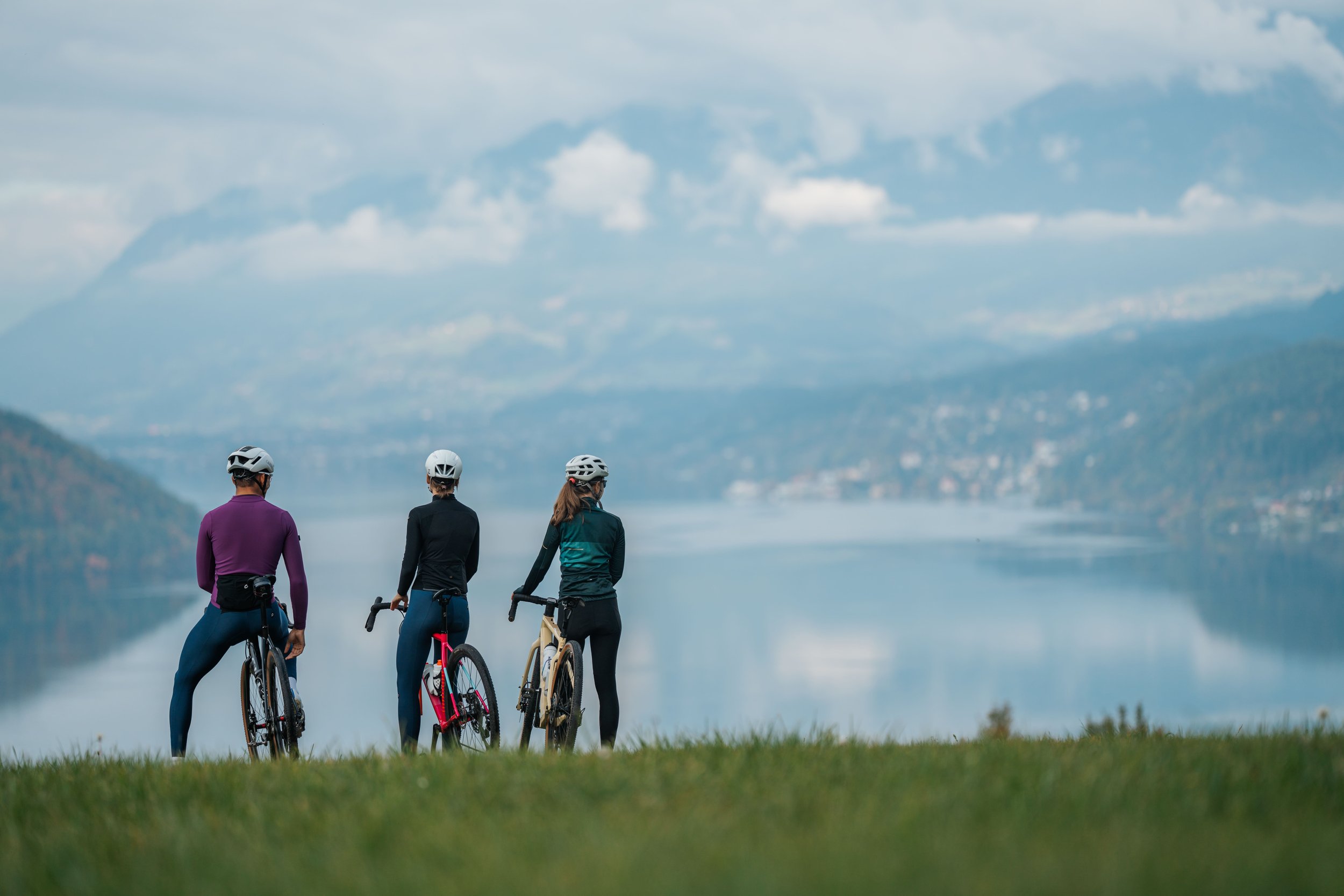 Drei Frauen mit Helmen fahren mit Fahrrädern an einem See entlang, im Hintergrund Berge und Wolken am Himmel.