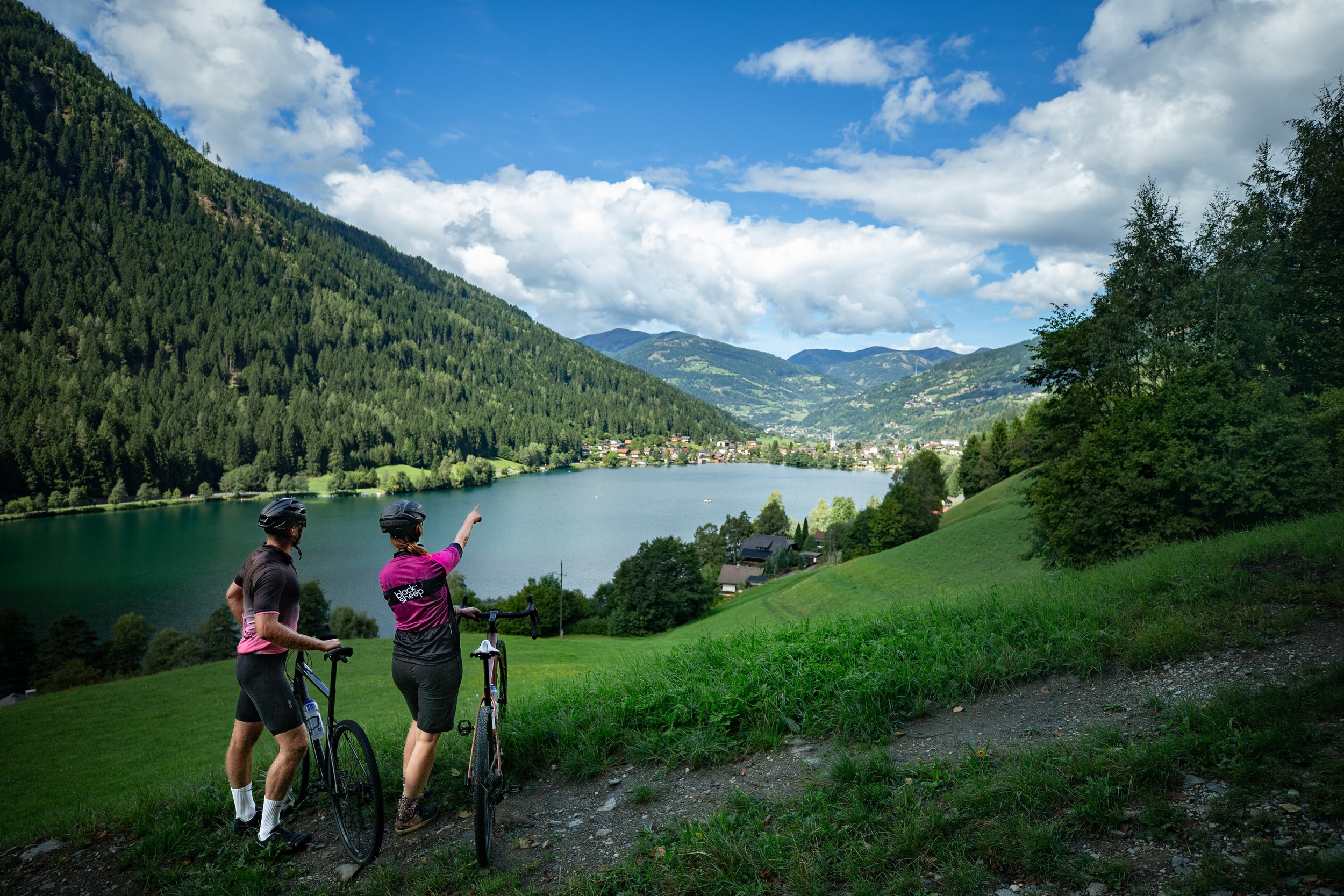 Zwei Radfahrer stehen auf einem grasbewachsenen Hügel, blicken auf einen See und Bergen. Ein Mann und eine Frau, beide mit Helmen, schauen und der Frau zeigt nach vorne. Es ist ein sonniger Tag mit einigen Wolken am Himmel.