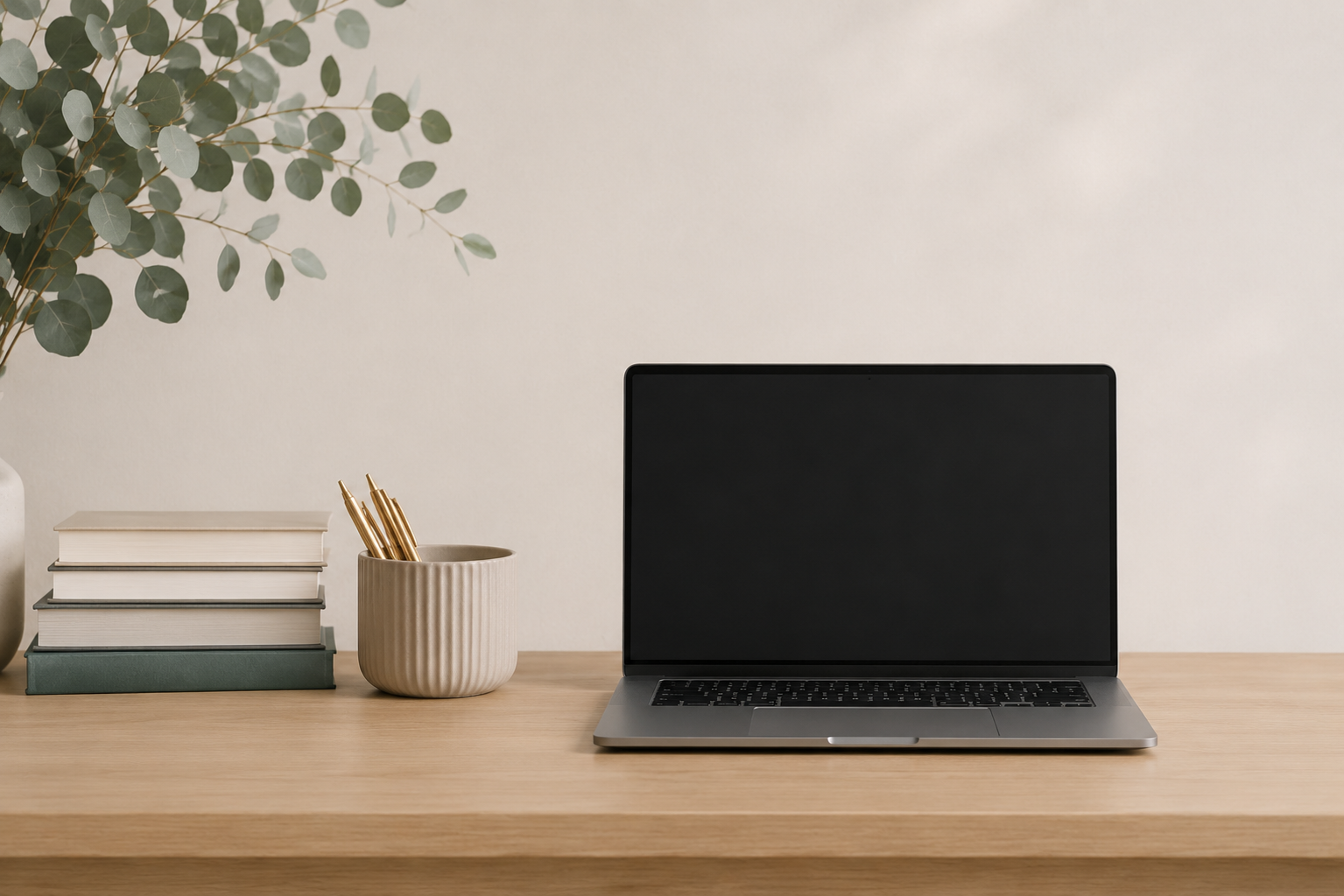 Minimalist workspace with a laptop, stacked books, a ceramic cup holding pens, and a leafy plant on a wooden desk against a plain wall.