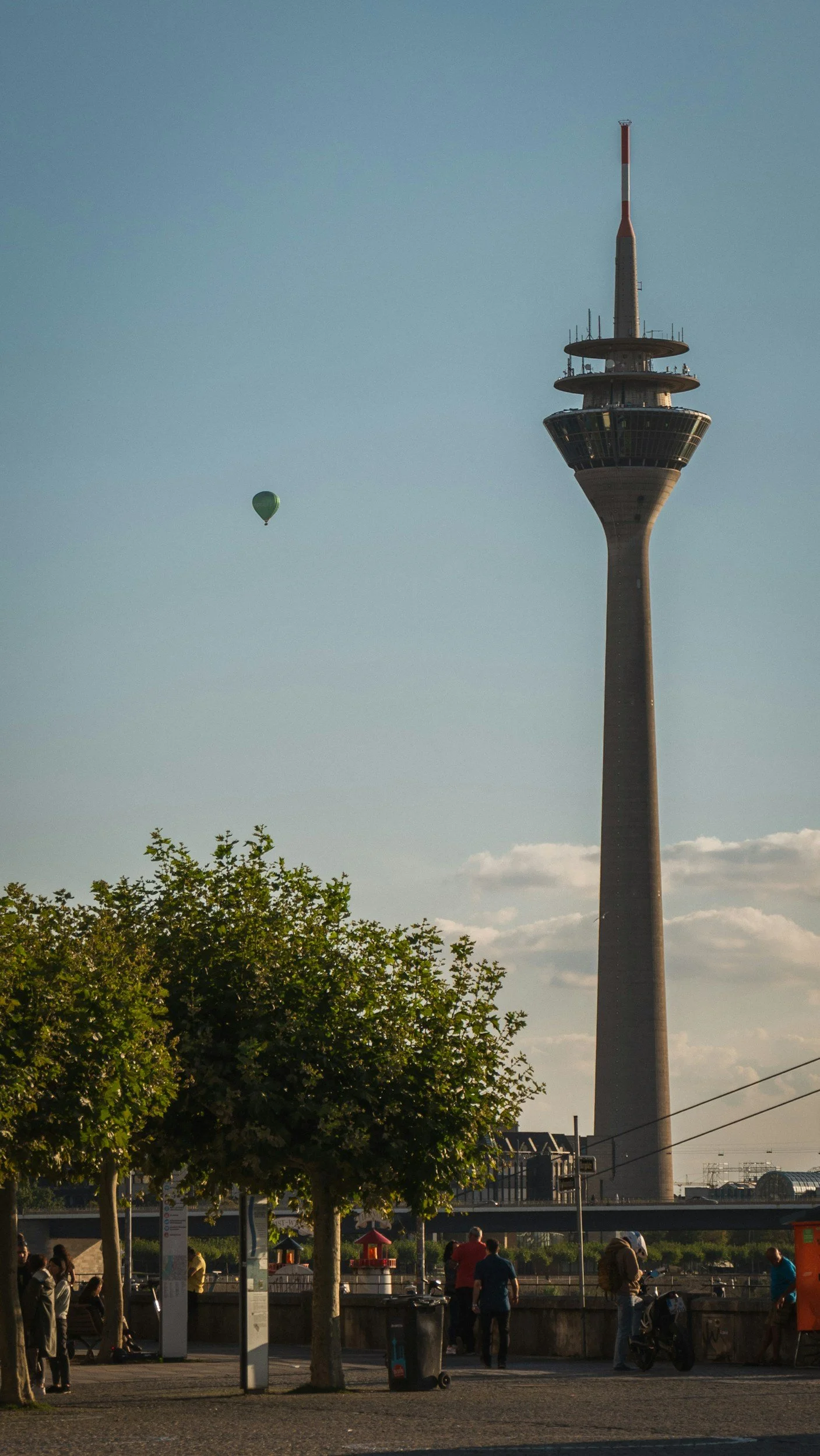 Stadtansicht mit Fernsehturm, Bäumen im Vordergrund, Menschen und Straßenbahn in einer urbanen Umgebung.