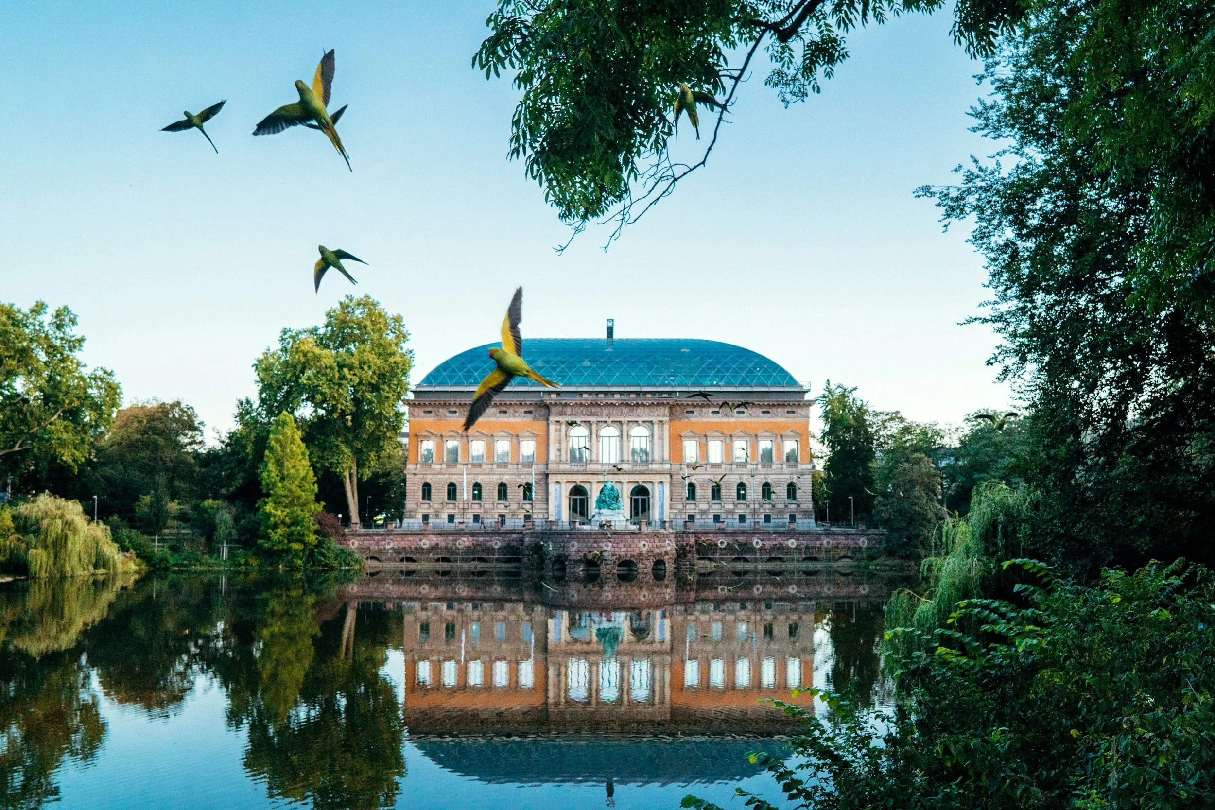 Historisches Gebäude mit blauer Kuppel, umgeben von Bäumen, Wasser reflektiert das Gebäude und die Natur, Hummeln fliegen vor Himmel.
