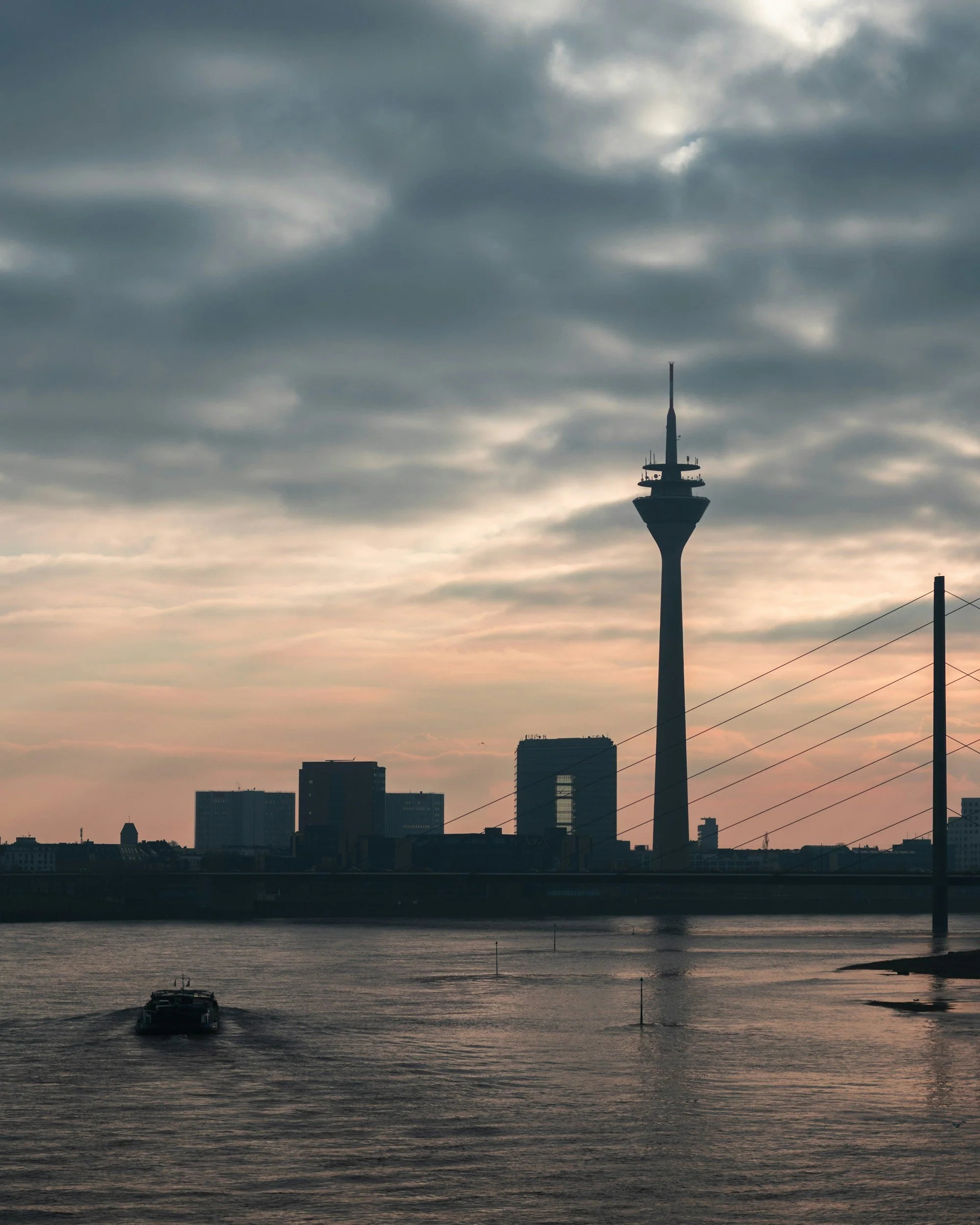Stadtbild mit Wasser, Wolkenhimmel, Fernsehturm im Vordergrund und Hochhäuser im Hintergrund bei Sonnenuntergang
