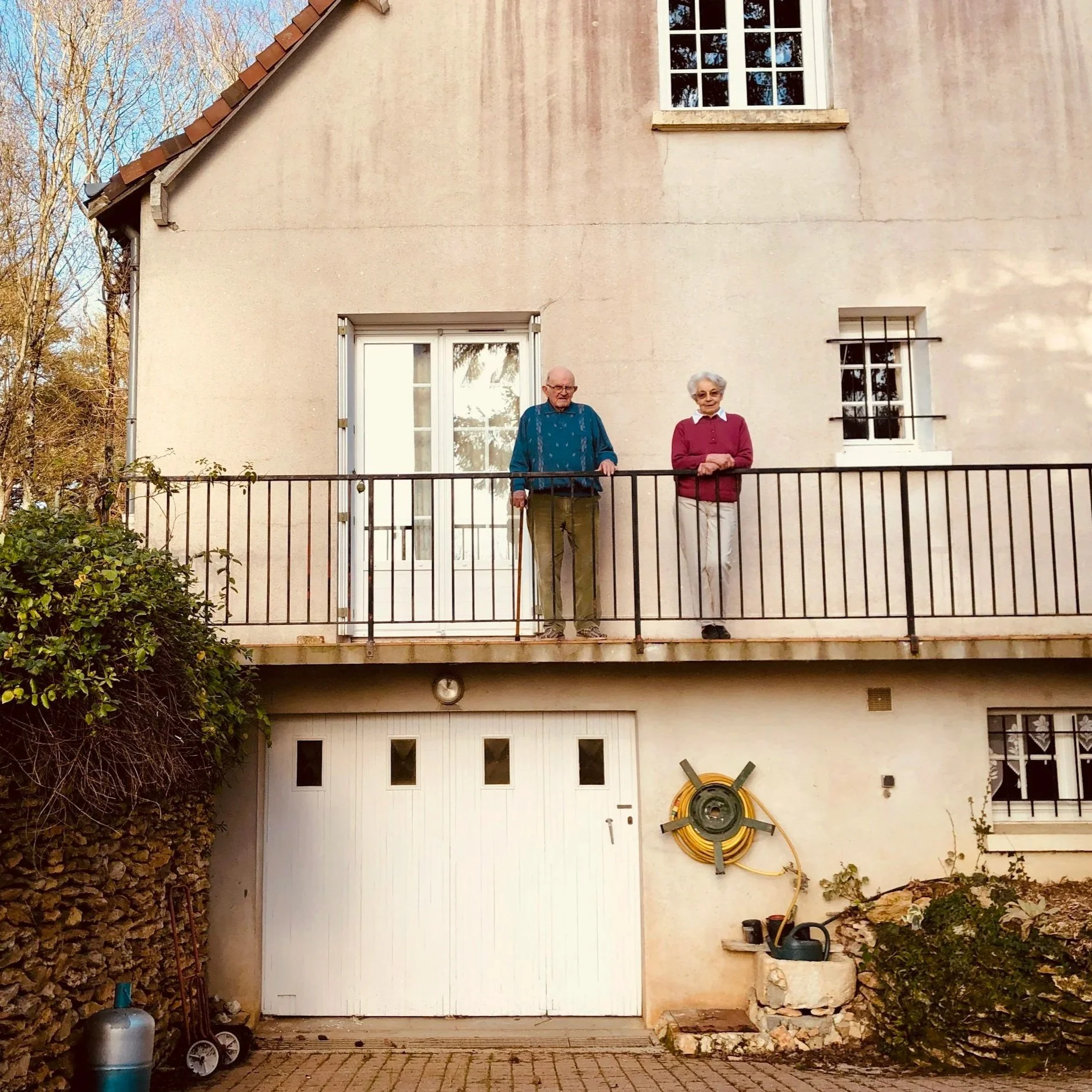 Ein zweistöckiges Haus mit einer weißen Tür und drei Fenstern, zwei im oberen Stockwerk und eines im unteren. Es gibt einen Balkon mit einem alten Mann und einer älteren Frau, die am Geländer stehen. Der Garten ist mit Steinen und Pflanzen gestaltet, auf der linken Seite ist eine Hecke, auf der rechten Seite befinden sich Gartengeräte und ein Wasserschlauch. Das Haus hat eine beige Fassade und ein Satteldach mit roten Ziegeln.
