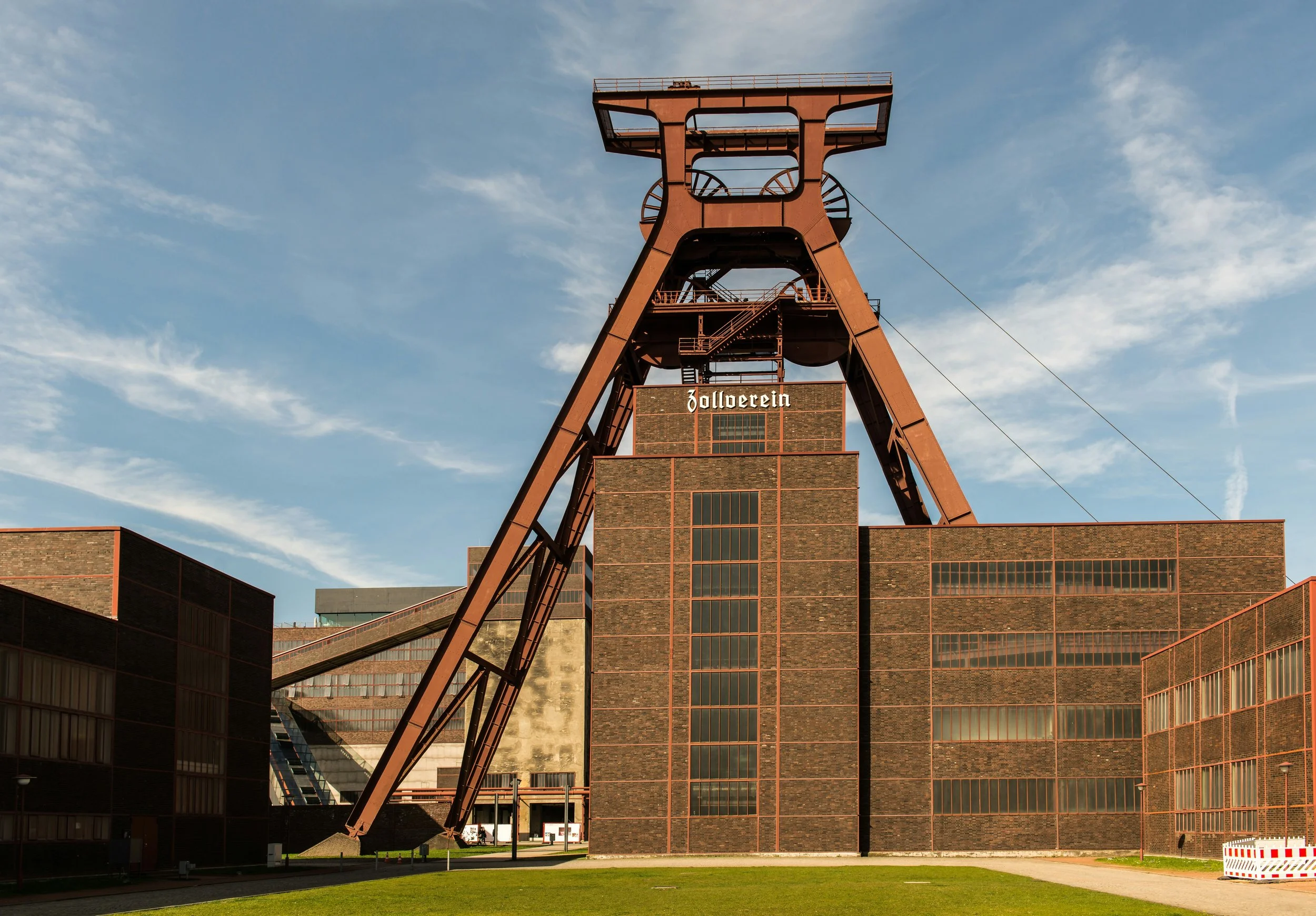 Ein ehemaliger Bergwerksförderturm aus rotem Stahl vor einem blauen Himmel mit Wolken, Teil eines Industriekomplexes mit modernen Gebäuden im Hintergrund.