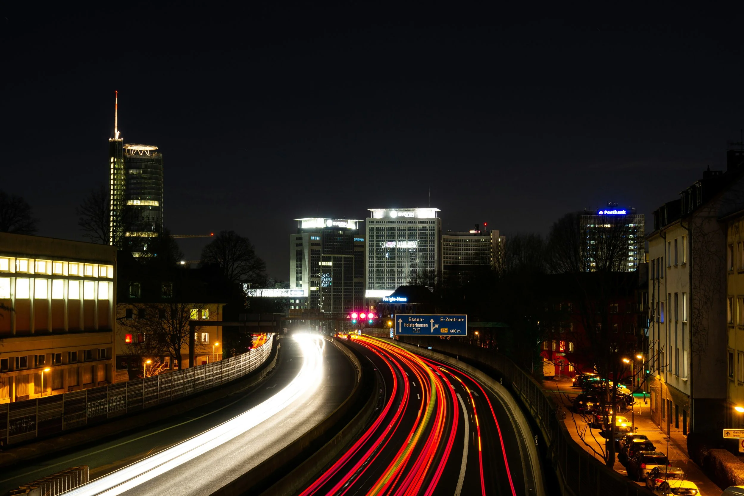Stadtansicht bei Nacht mit beleuchteten Hochhäusern, Straßenverkehr mit Lichtspuren, Straßen und Stadtverkehr, Verkehrszeichen, Häuser und Bäume im Vordergrund.
