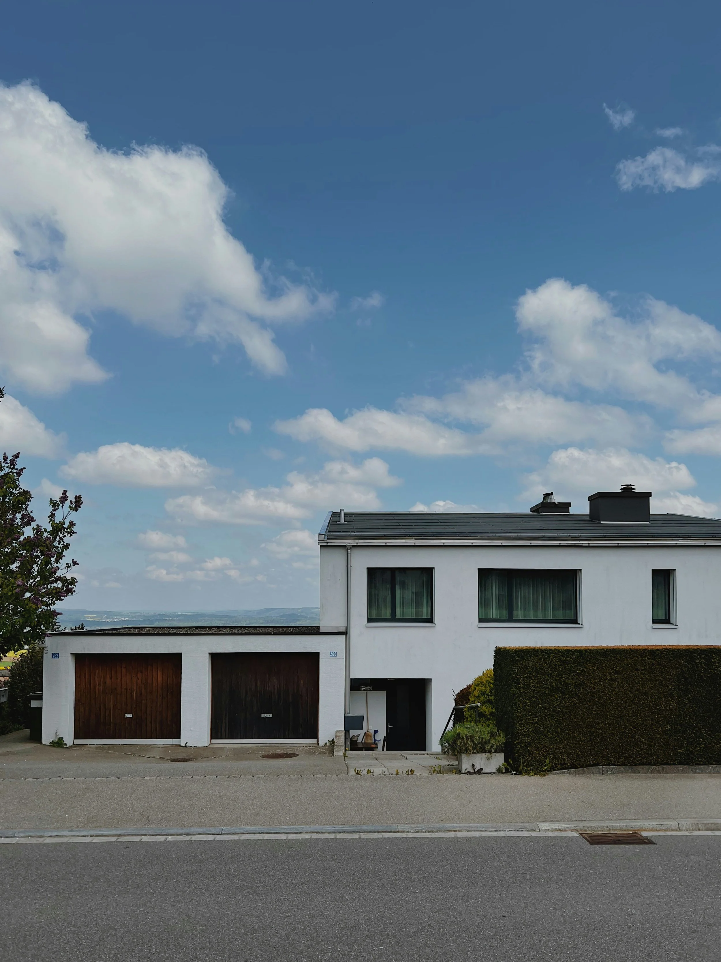 Moderner weißer Wohnhaus mit zwei oberen Fenstern, Parkplatz und grüner Hecke vor der Straße, blaue Himmel mit weißen Wolken im Hintergrund.