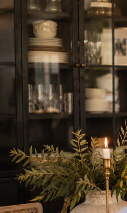 A dark wooden cabinet with glass doors displaying white and clear dinnerware and glassware, with a leafy green plant and a lit candle in the foreground.