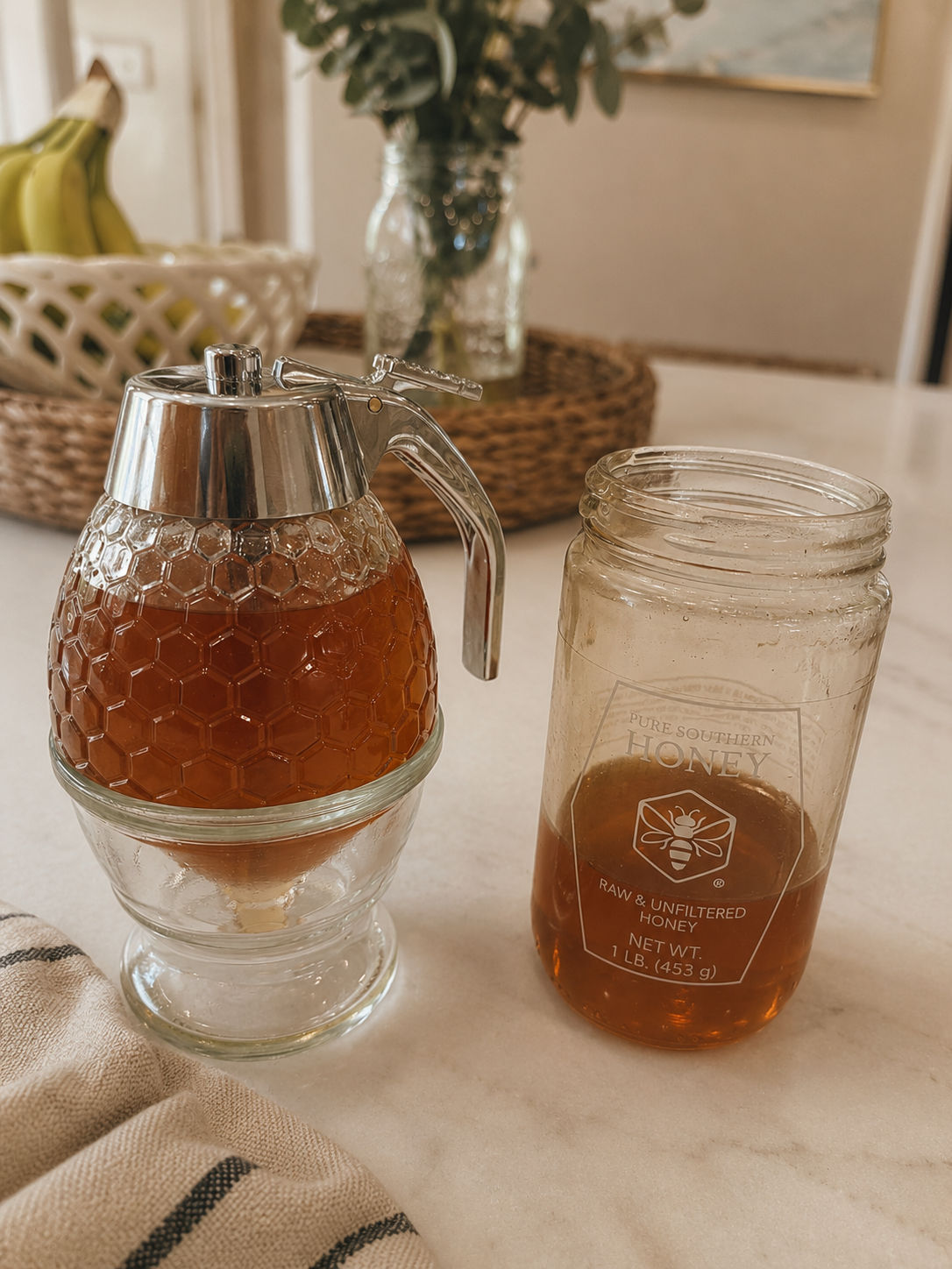 A glass honey dispenser filled with honey and a glass jar of honey with a label reading 'Pure Southern Honey, Raw & Unfiltered.'