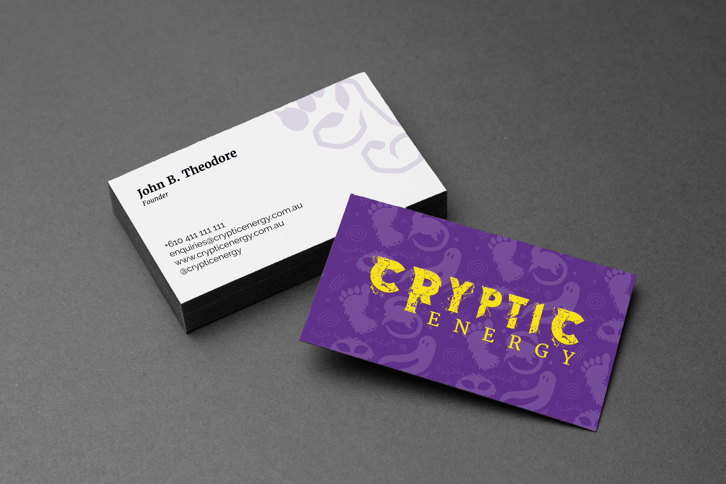 Stack of business cards on a gray surface, one card partially visible with purple and yellow text and design related to cryptic energy, and another white card with contact information for John B. Theodore.