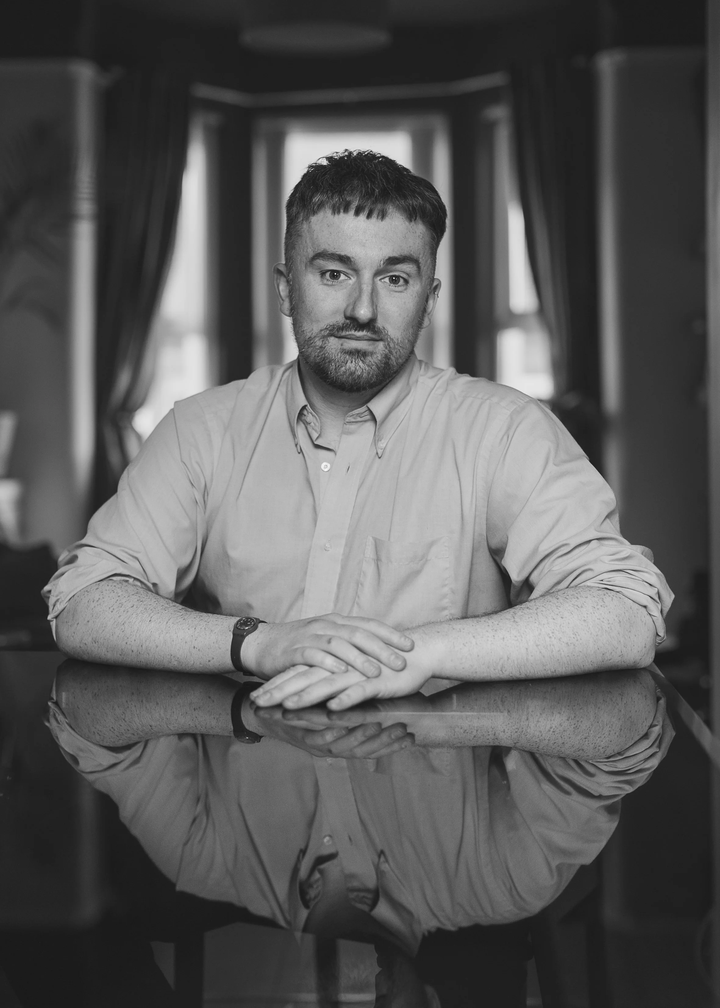 A black and white photo of a man with short hair and a beard, sitting at a table with his arms crossed on the surface. There is a window behind him, framed by curtains, in a room with dim lighting.