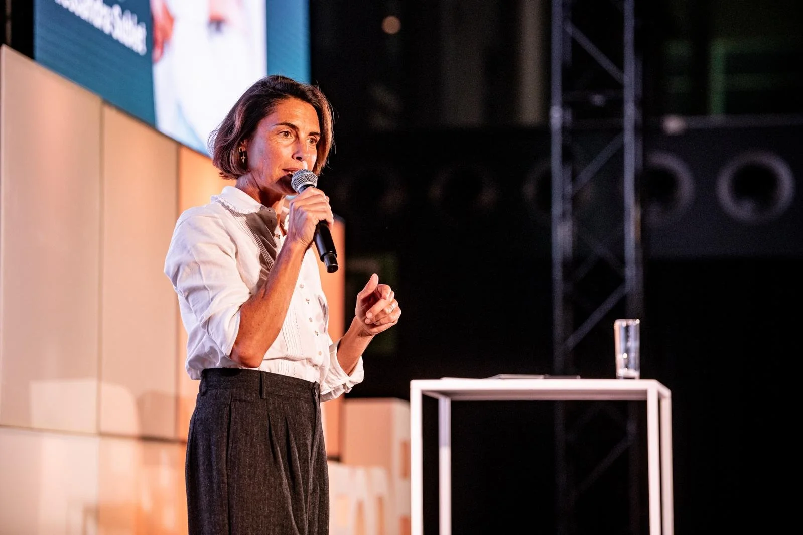 Femme debout parlant dans un microphone lors d'un événement, portant une chemise blanche et un pantalon sombre, avec une petite table à côté et un écran en arrière-plan.