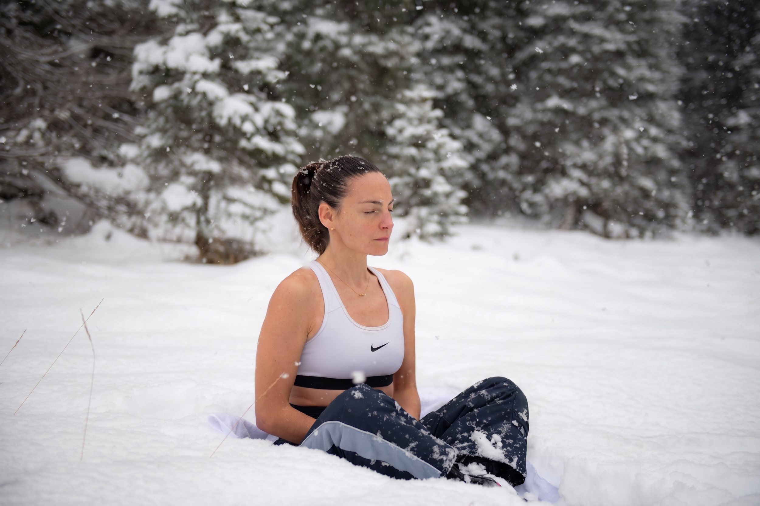 Une femme en tenue de sport blanche et noire méditant en tailleur dans la neige, avec des arbres enneigés en arrière-plan.