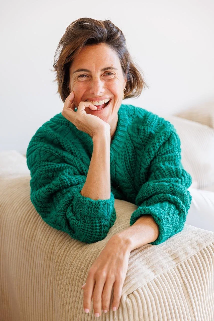 Une femme souriante, portant un pull vert, assise sur un canapé beige, dans une pièce lumineuse.