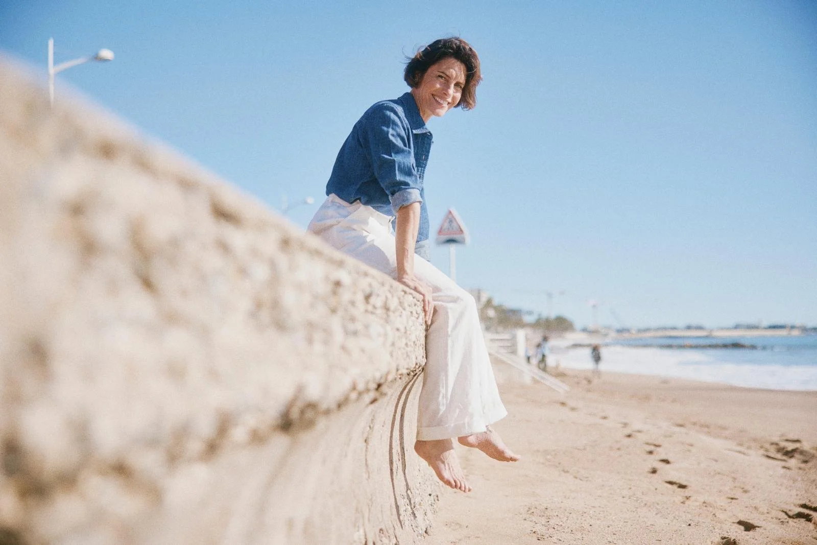 Femme souriante assise sur une barrière en béton au bord de la plage, pieds dans le sable, avec la mer en arrière-plan, ciel clair et soleil brillant.