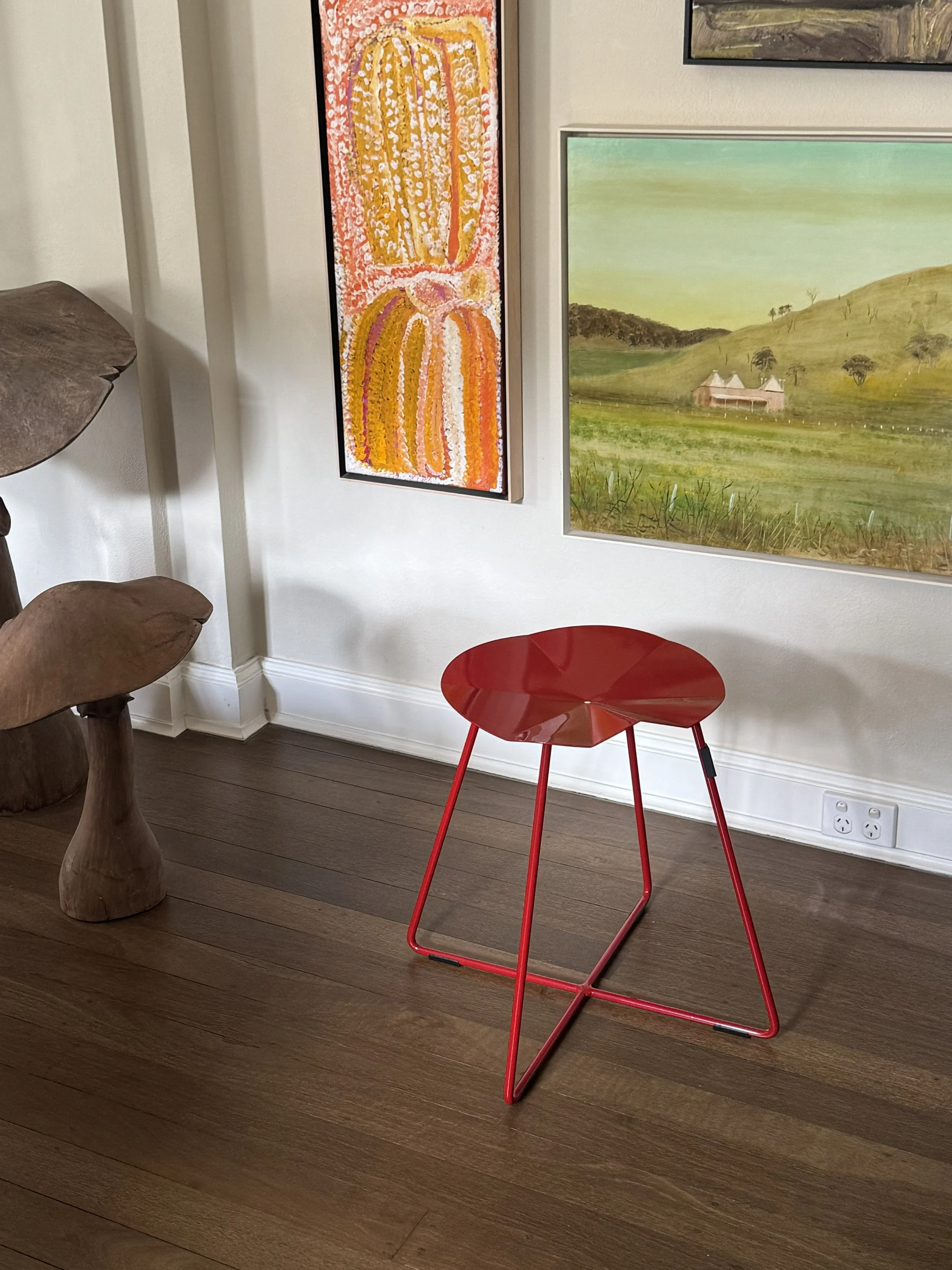 Red modern chair with thin metal legs on wooden floor in front of a white wall with artwork and a power outlet