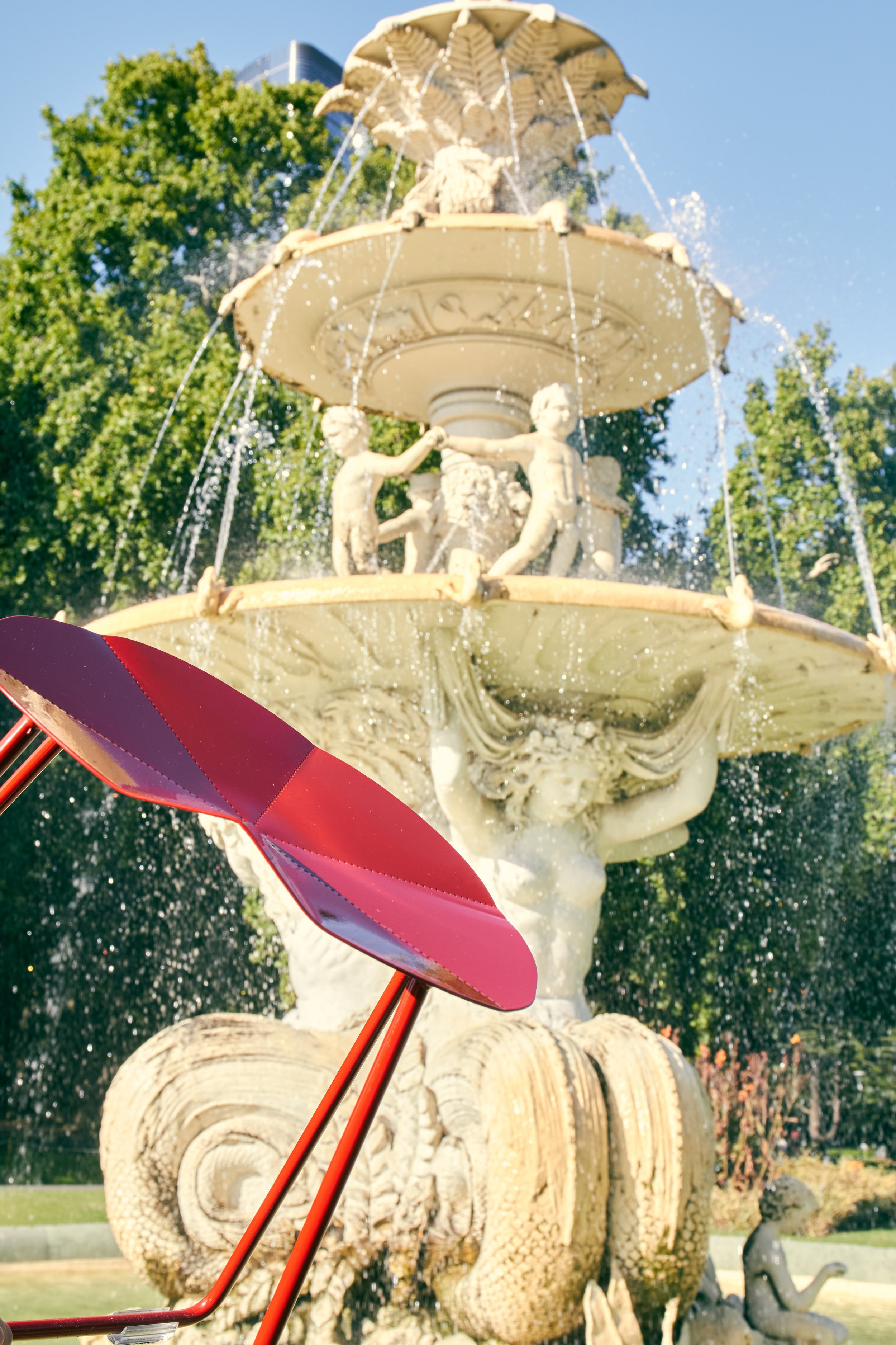 A view of a decorative water fountain featuring four fish sculptures spewing water, with trees and a building in the background, and a purple and red umbrella in the foreground.