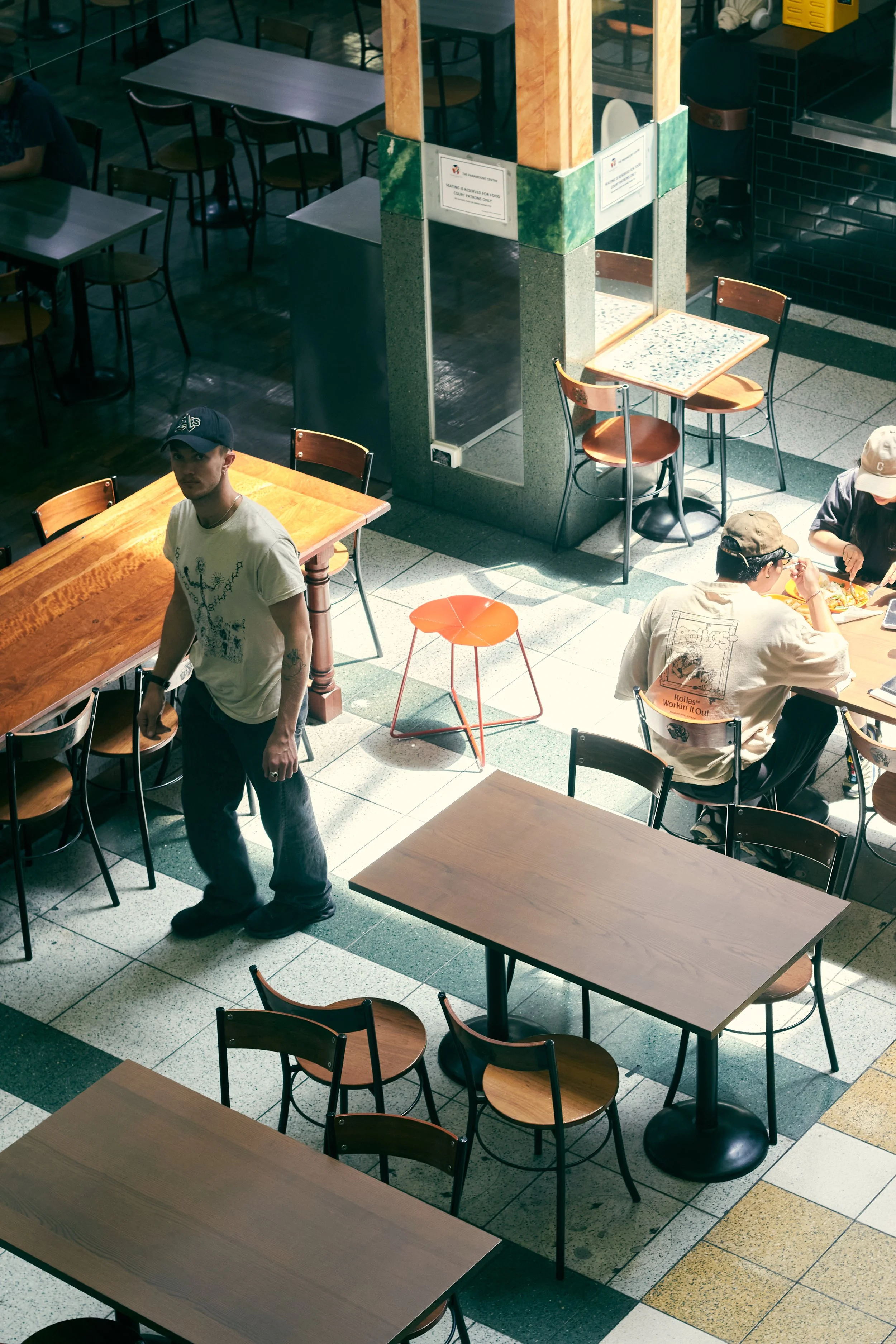 Interior of a dining area with empty wooden tables and chairs, some tables are occupied with people eating, and a man standing near a table. The space has a tiled floor and a decorated central pillar with signs.