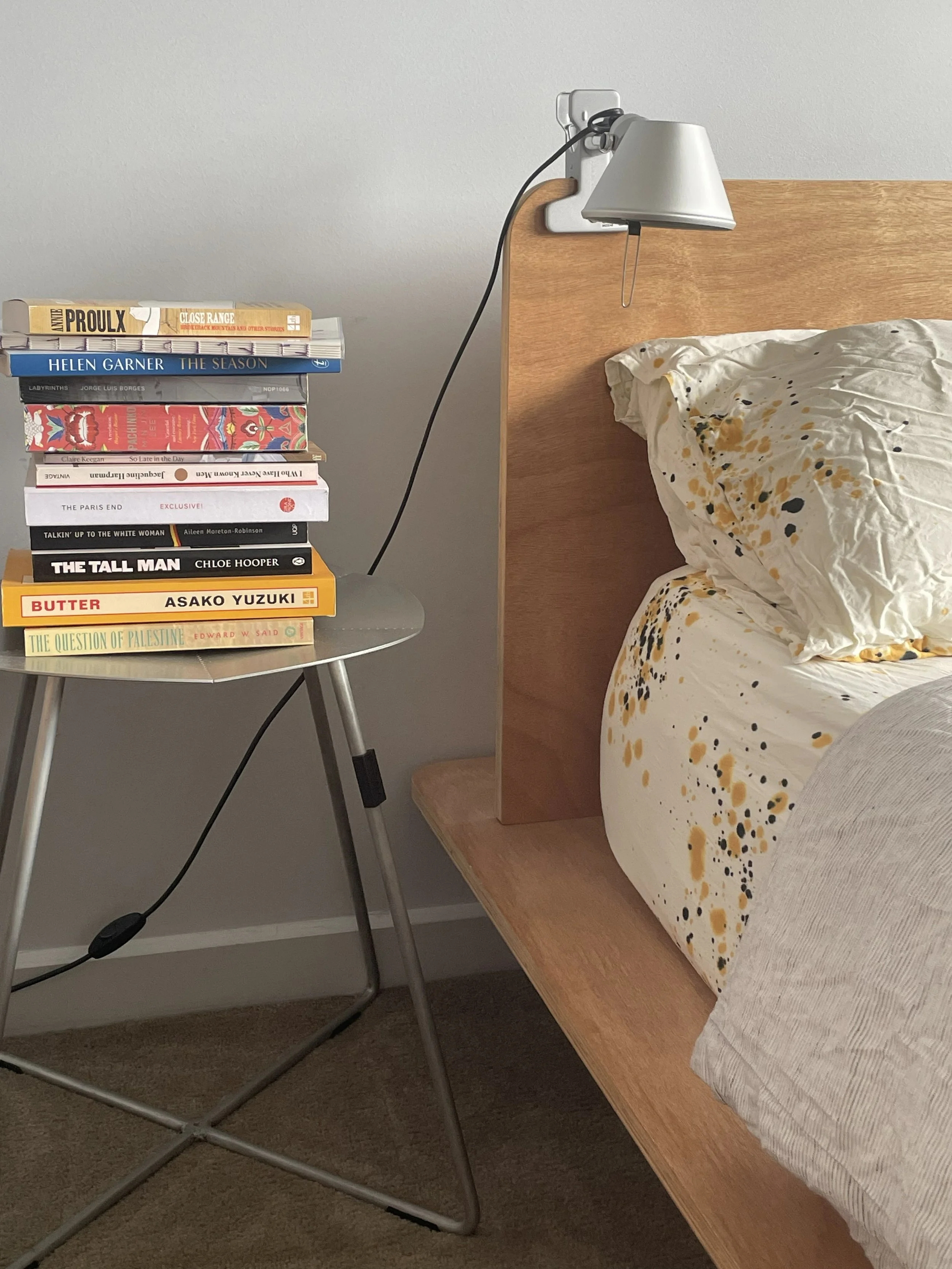 A bedside table with a stack of books next to a bed with white, yellow, and black splattered bedding. There is a wall-mounted lamp attached to a wooden headboard.