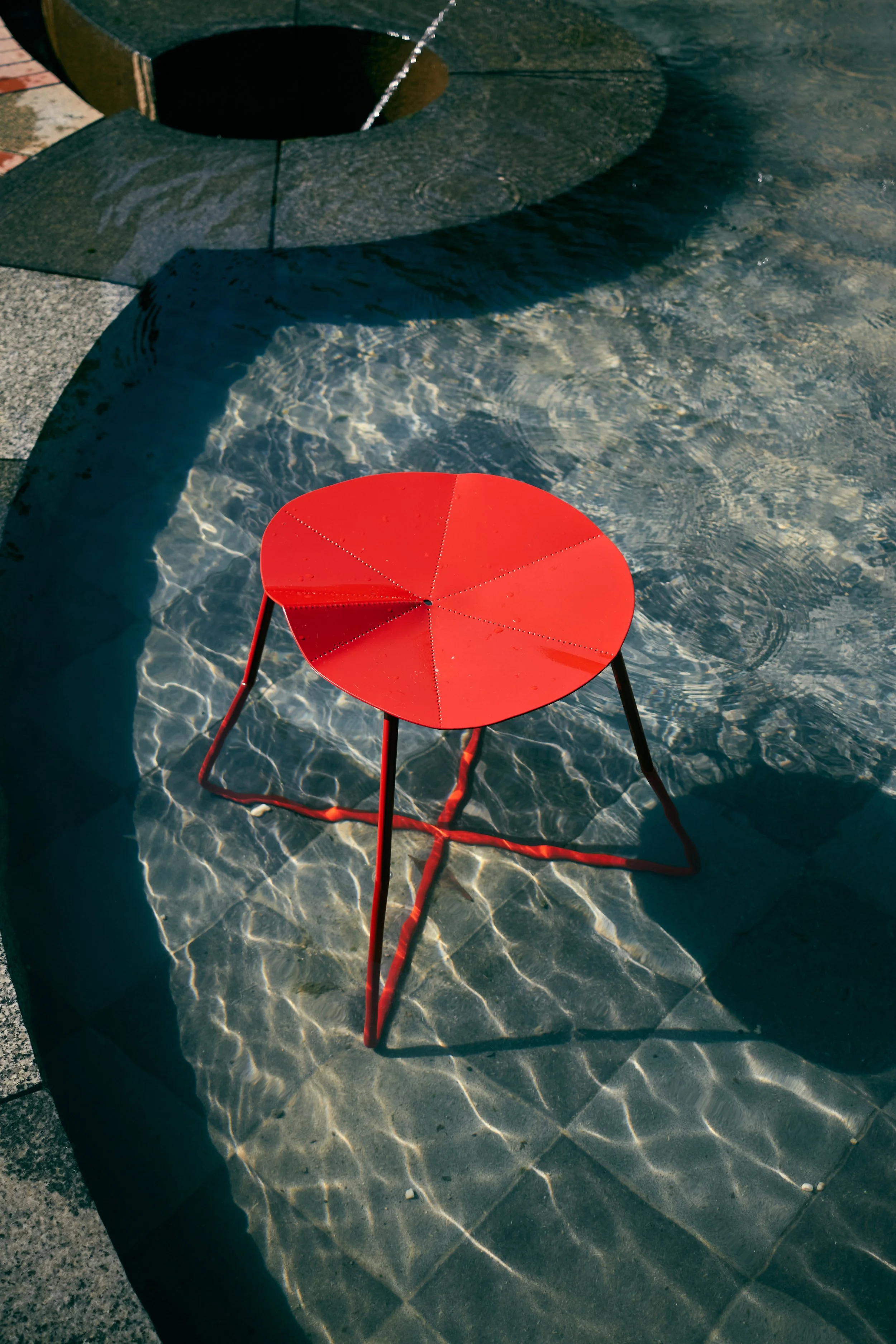 Red round metal patio table with four legs, sitting on a concrete ground next to a pool with a stone border, with sunlight creating water ripple reflections on the pool surface.