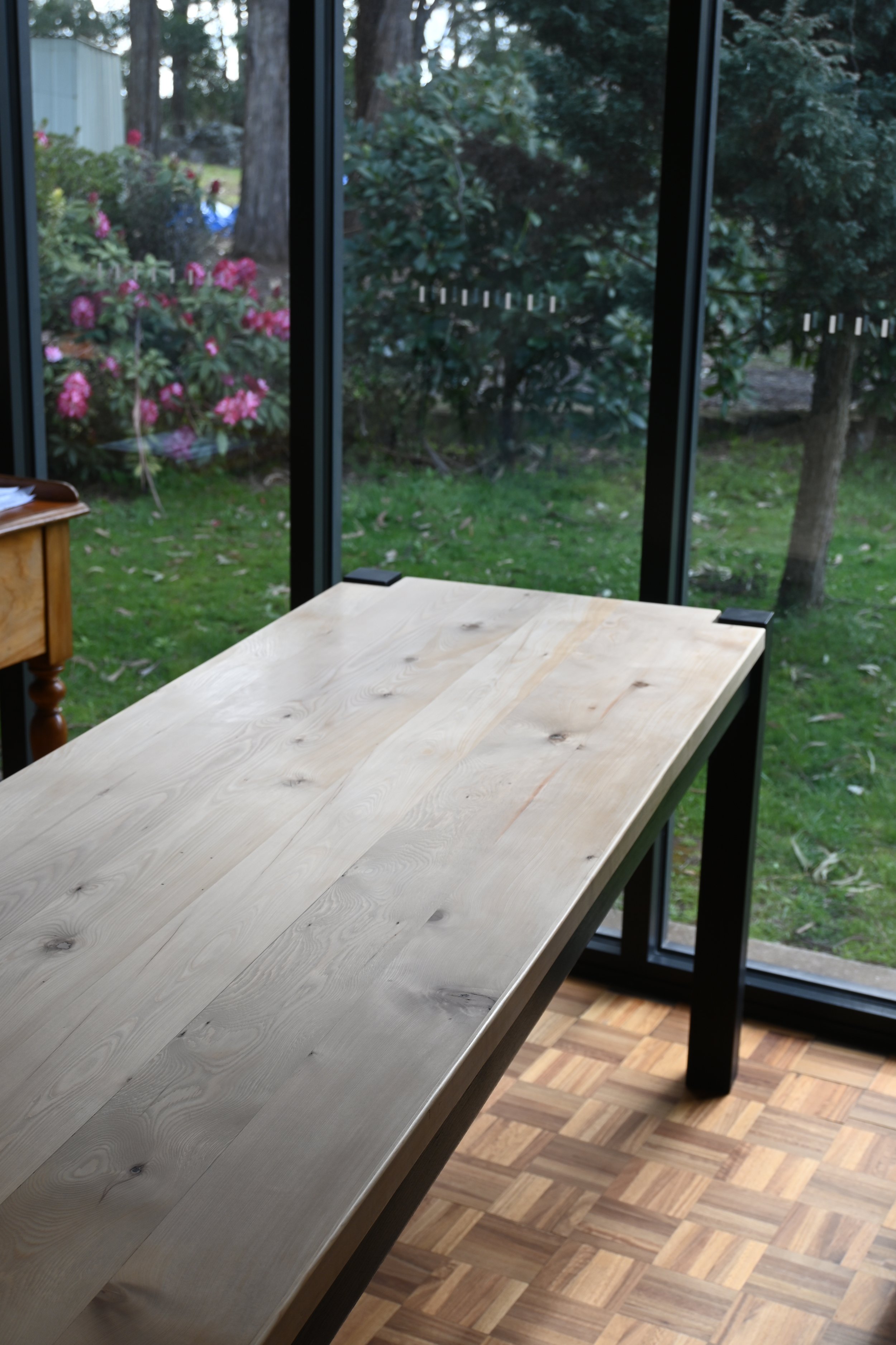 A partially assembled light-colored wooden table inside a room, with a backyard visible through large glass sliding doors, blooming pink flowers, green grass, and trees outside.