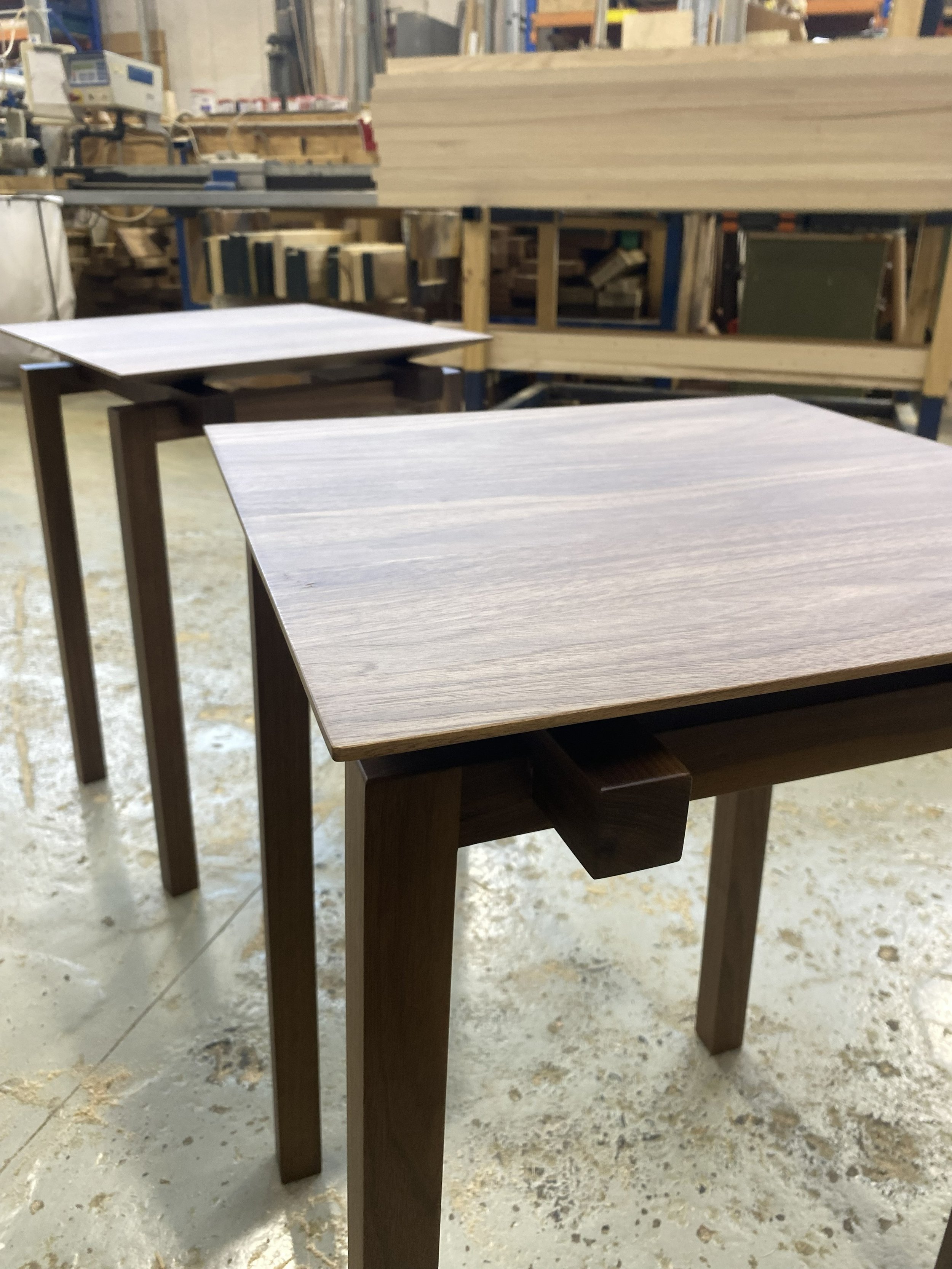 Two wooden tables with dark brown finishes in a workshop, with a larger table in the background. The workshop has shelves and tools visible, and the floor is concrete with wood shavings or dust.