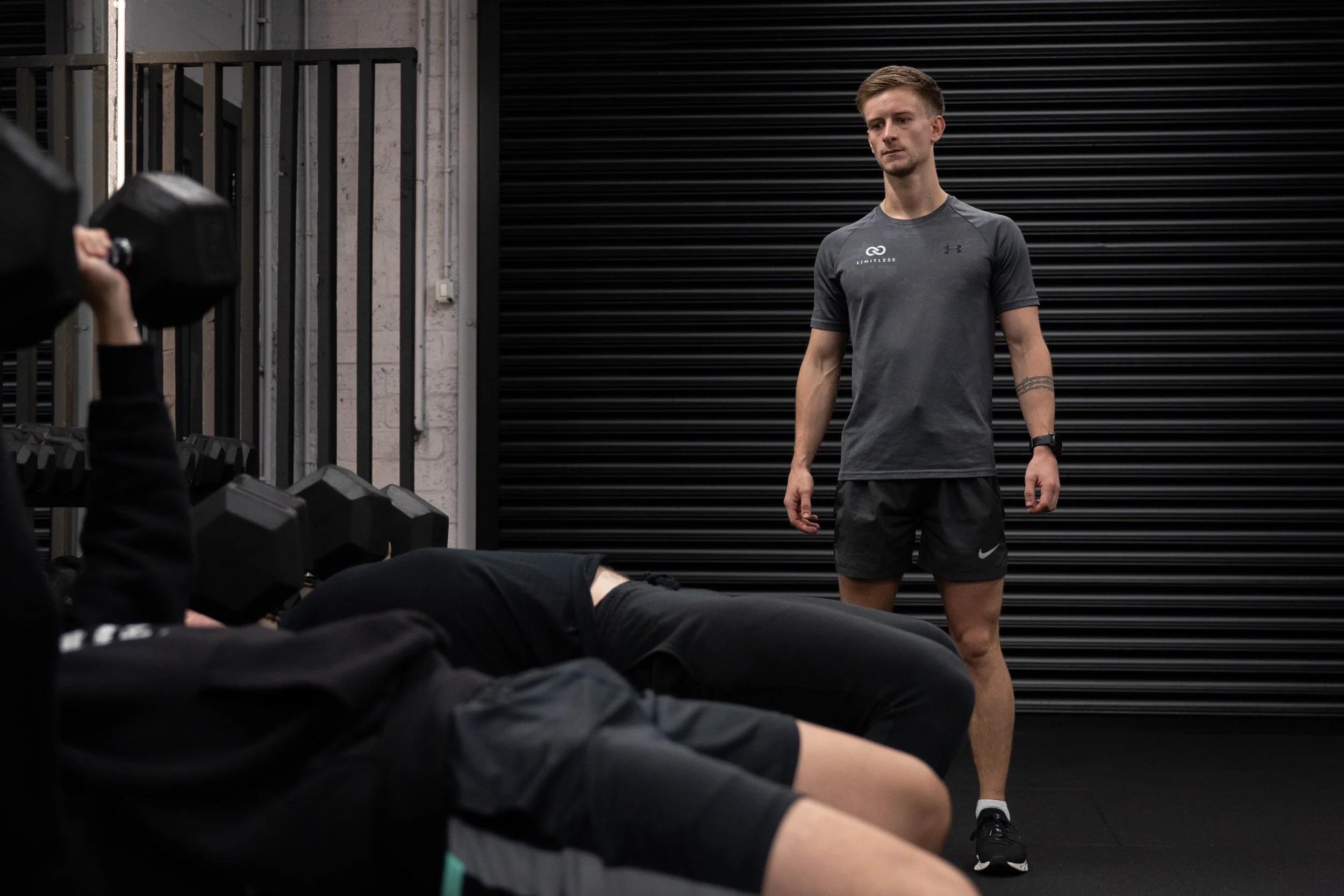 A man in a gray athletic shirt and black shorts standing in front of a group sitting on the gym floor, with a black industrial door behind him.