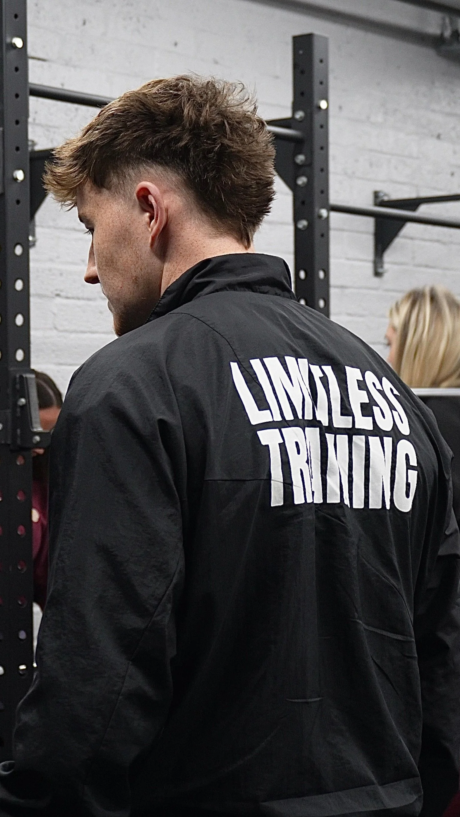 A young man with a messy hairstyle wearing a black jacket with the words 'Limitless Training' printed on the back, standing inside a gym with weightlifting equipment and other people in the background.
