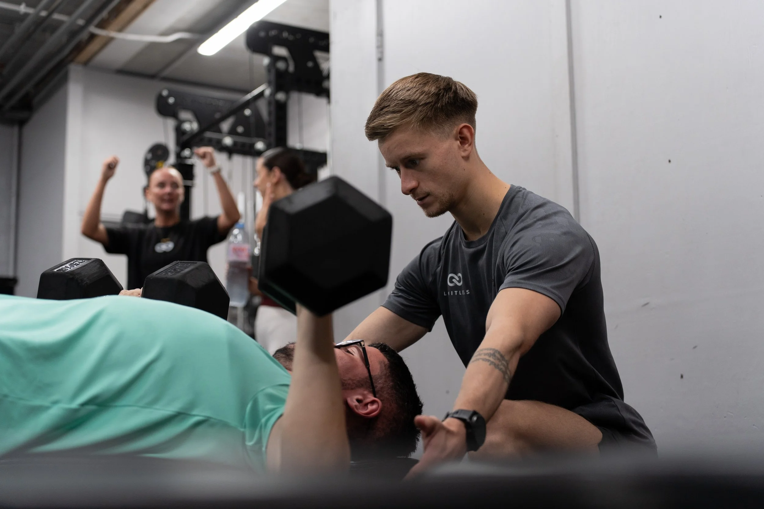 A man in a black shirt assisting a man in a green shirt with an exercise at a gym. Two women are in the background, celebrating or cheering.