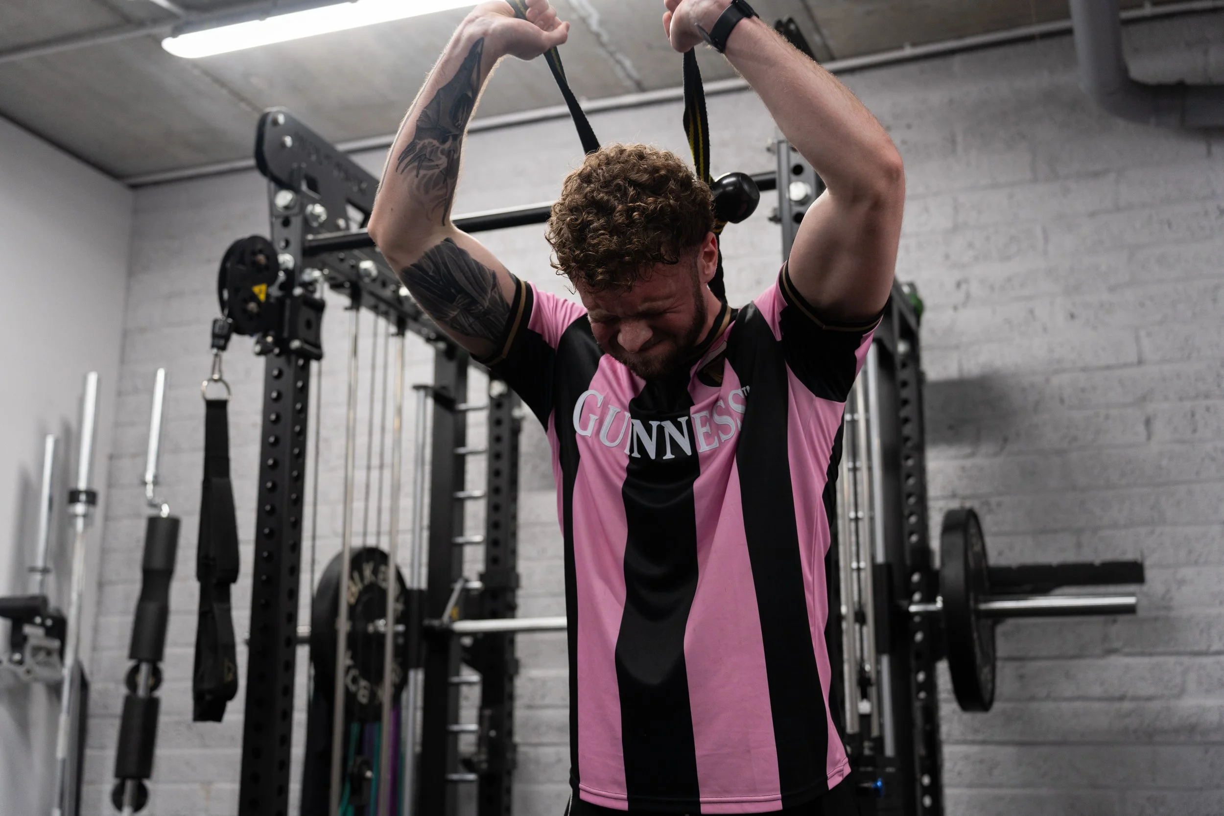 A man with curly hair and tattoos on his arm is lifting a black and yellow suspension trainer in a gym, wearing a pink and black sports jersey with 'Guinness' written on it.