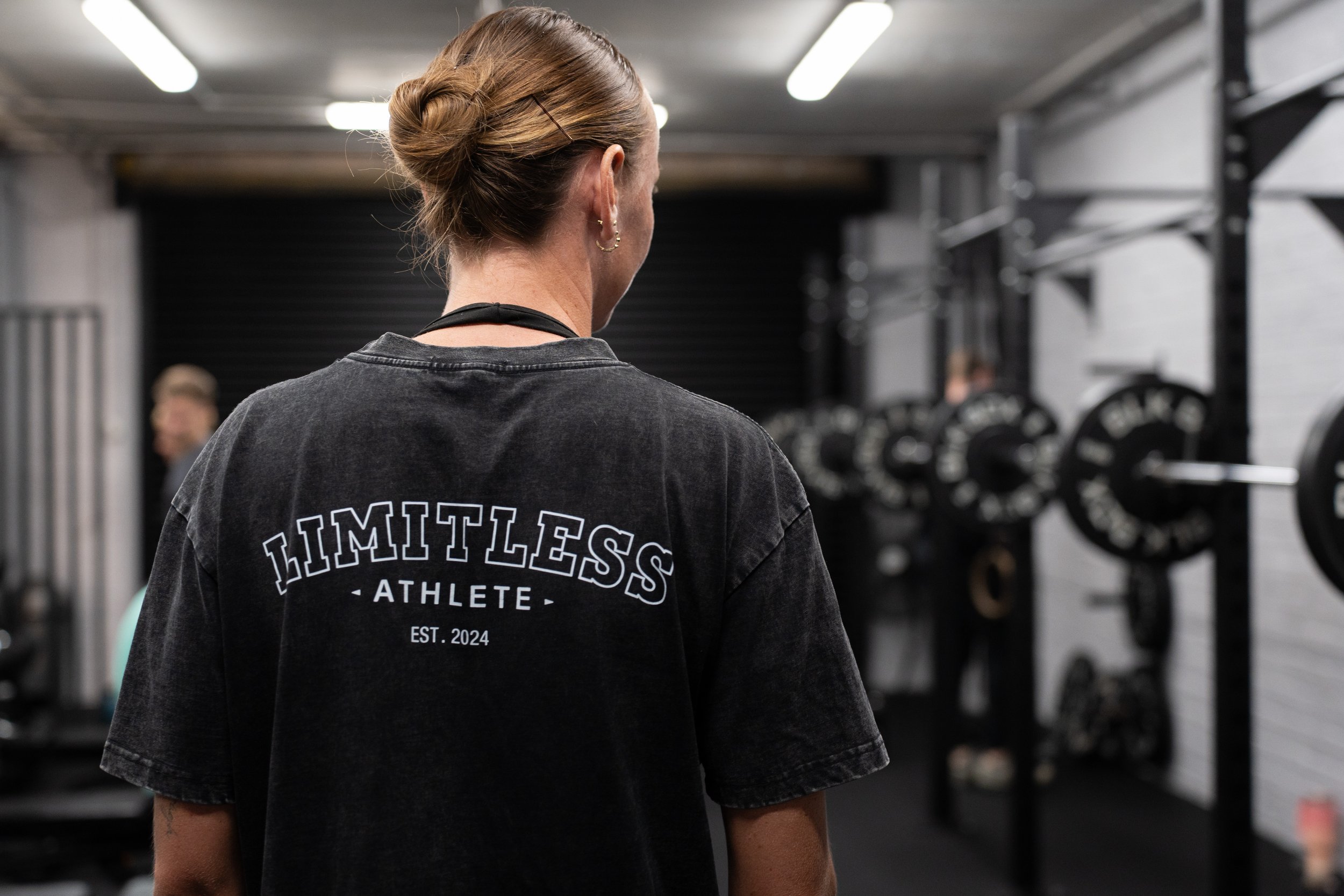 Back view of a woman in a gym wearing a black T-shirt with the text 'LIMITLESS ATHLETE EST. 2024'. She has her hair in a bun and is looking towards weightlifting equipment with people in the background.