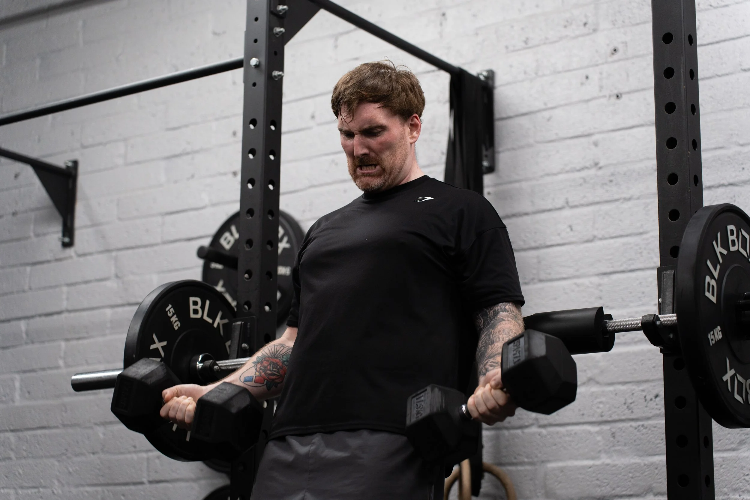 A man with tattoos on his arms lifting dumbbells in a gym, with a chain wall and black metal equipment in the background.