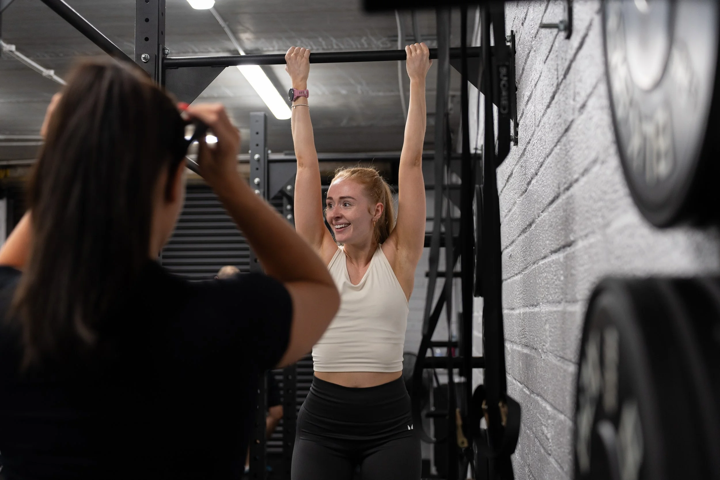 A woman with a red ponytail and a white tank top is doing pull-ups on a pull-up bar at the gym, smiling. Another person with long dark hair is taking a picture or video of her.