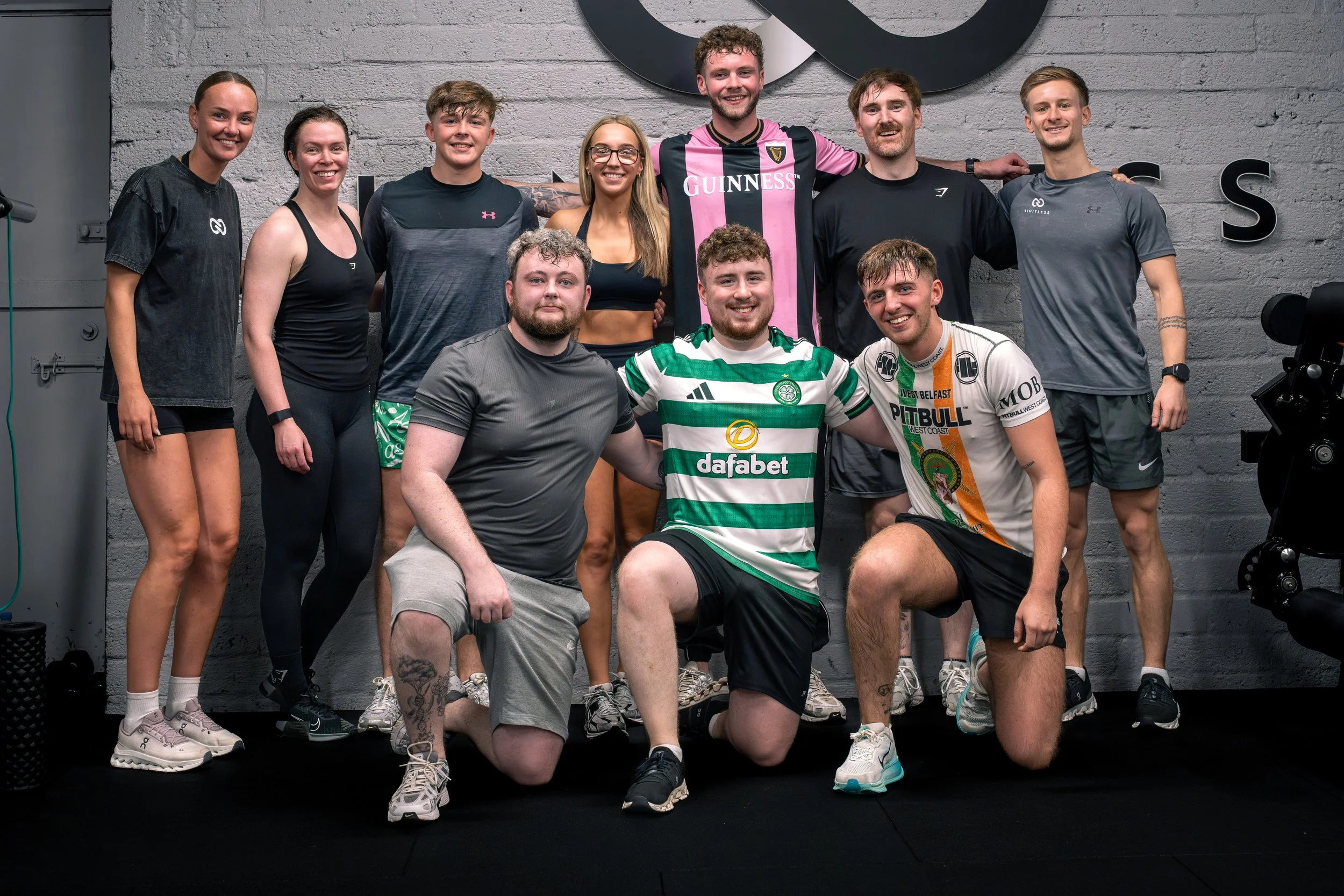 Group of ten fitness enthusiasts posing together in a gym, with some kneeling in the front row and others standing in the back row, in front of a gray brick wall.