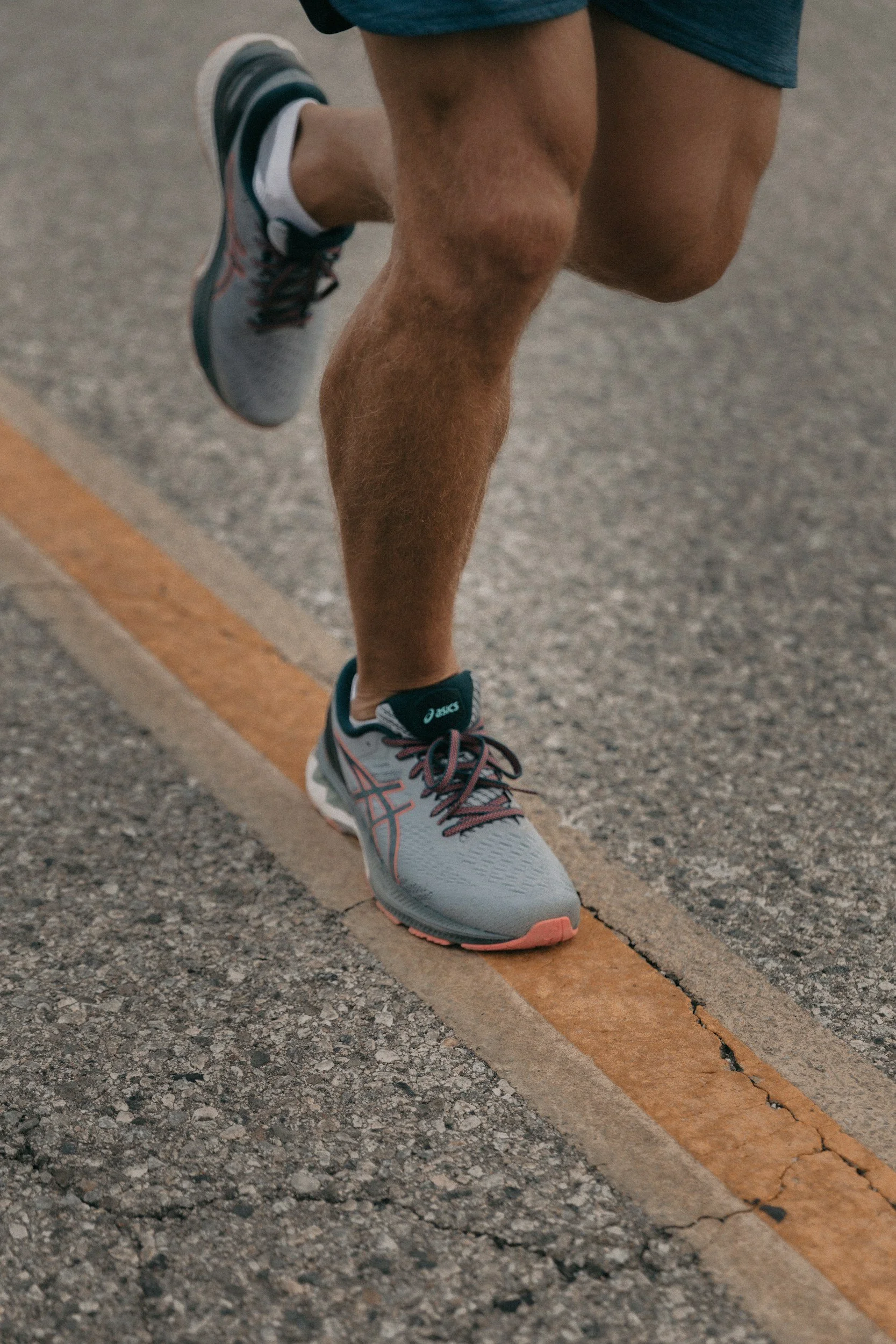 Une personne en train de courir sur une route en enlevant un pied, portant des chaussures de course gris avec des détails roses et noirs, des shorts bleus, et une jambe musclée.