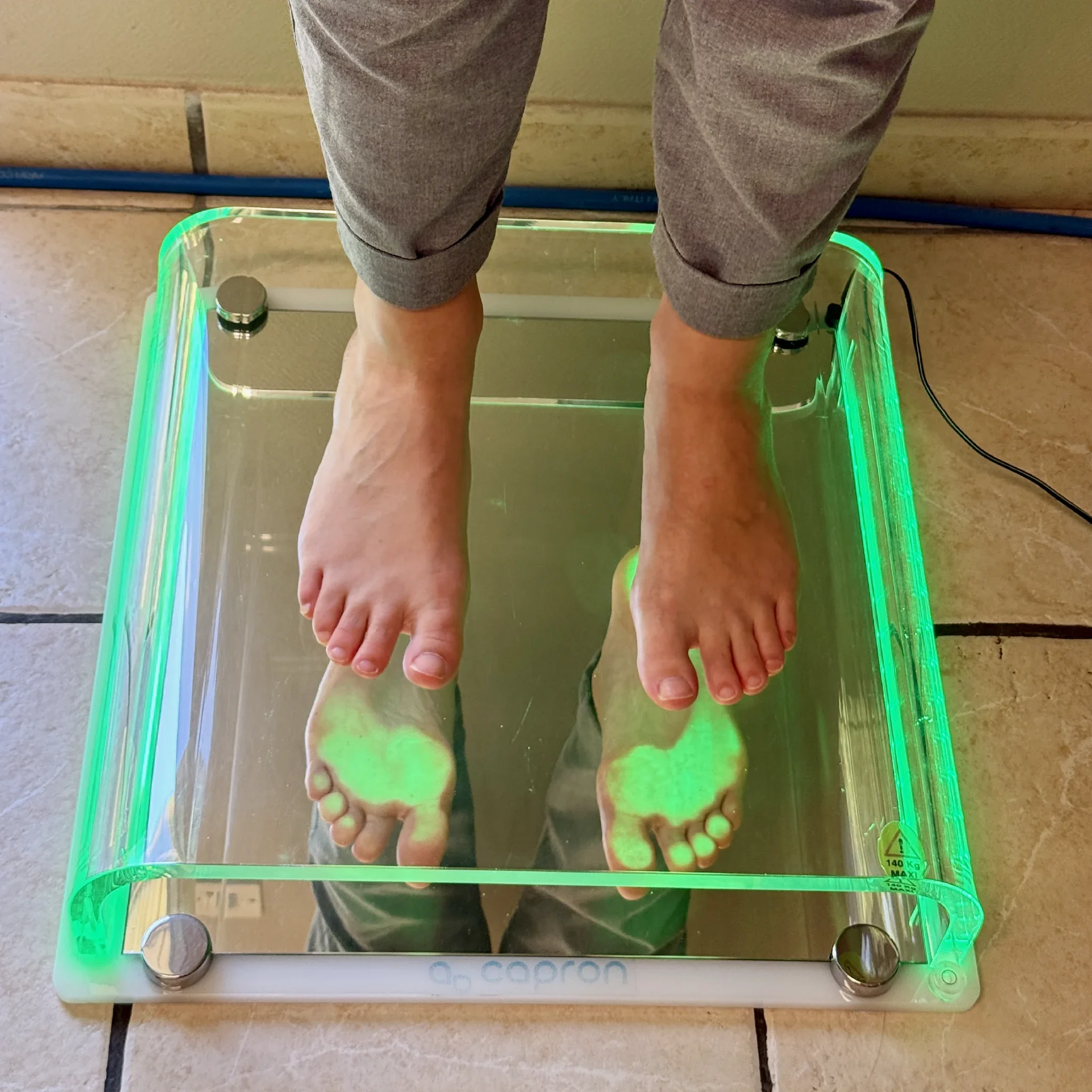 Une personne se tient sur une balance à minceur numérique avec un écran en verre calciné éclairé de lumières colorées, vue du dessus, montrant ses jambes et ses mains.
