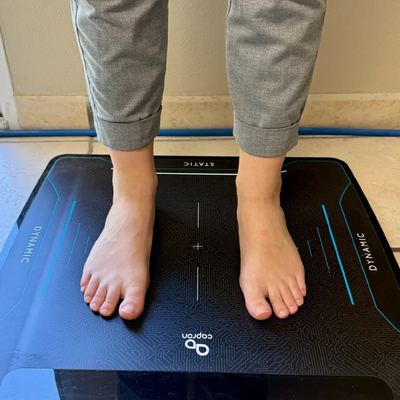 Pieds d'une personne debout sur une plateforme d'évaluation de la stabilité, avec des jambes en pantalon gris, dans une salle intérieure.