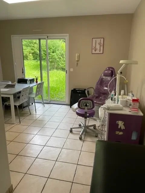 Salle de consultation équipée d'un fauteuil médical violet, d'une petite table avec des fournitures, d'une table avec un ordinateur, et une porte-fenêtre donnant sur un jardin vert.