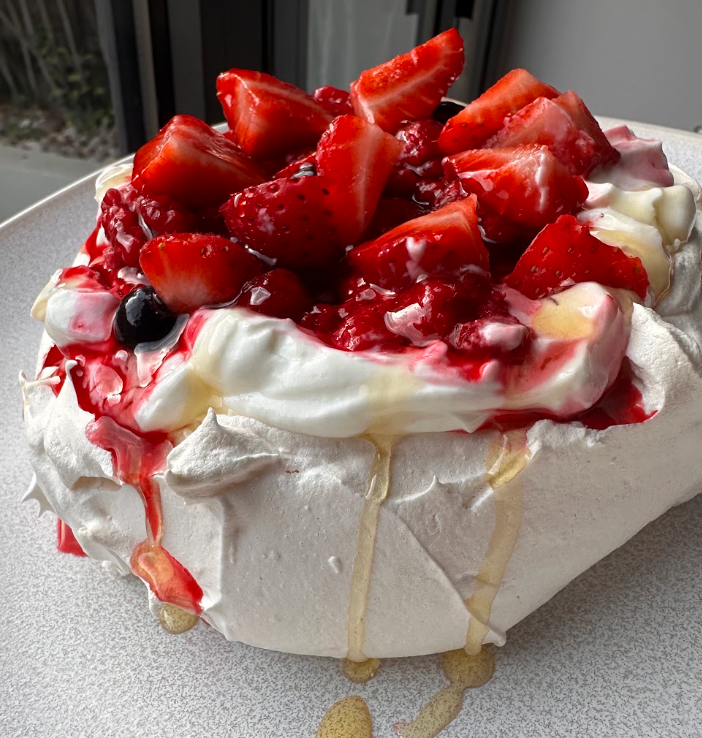 A dessert featuring meringue topped with whipped cream and mixed fresh berries, with berry syrup drizzled on top.
