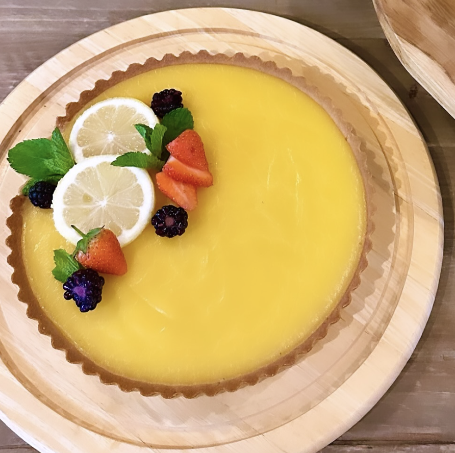 Lemon tart garnished with sliced lemon, strawberries, blackberries, mint leaves, and blackberries, on a round wooden serving tray.