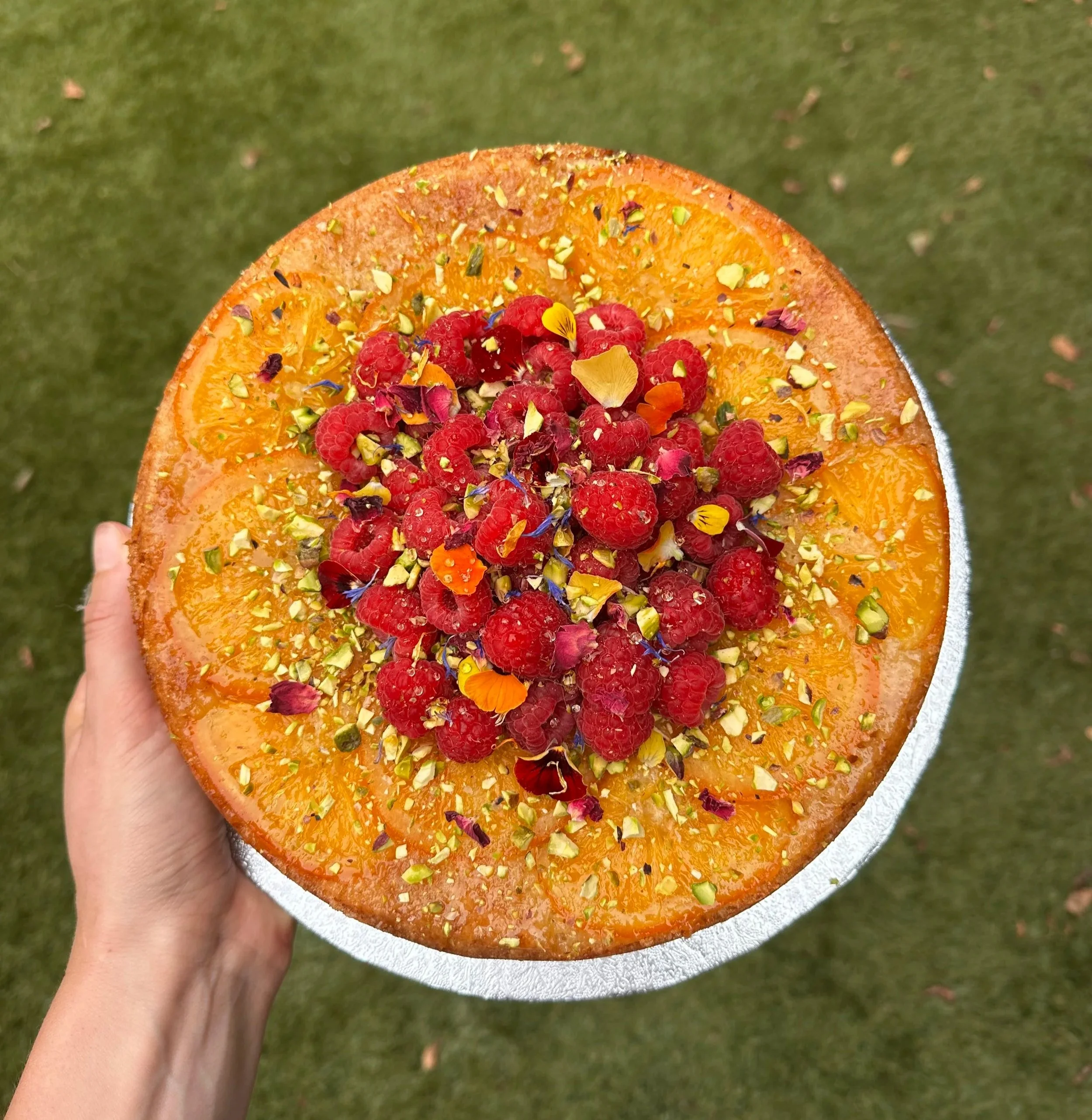 Orange, honey and almond cake with fresh raspberries, orange slices, pistachios, and edible flower petals.
