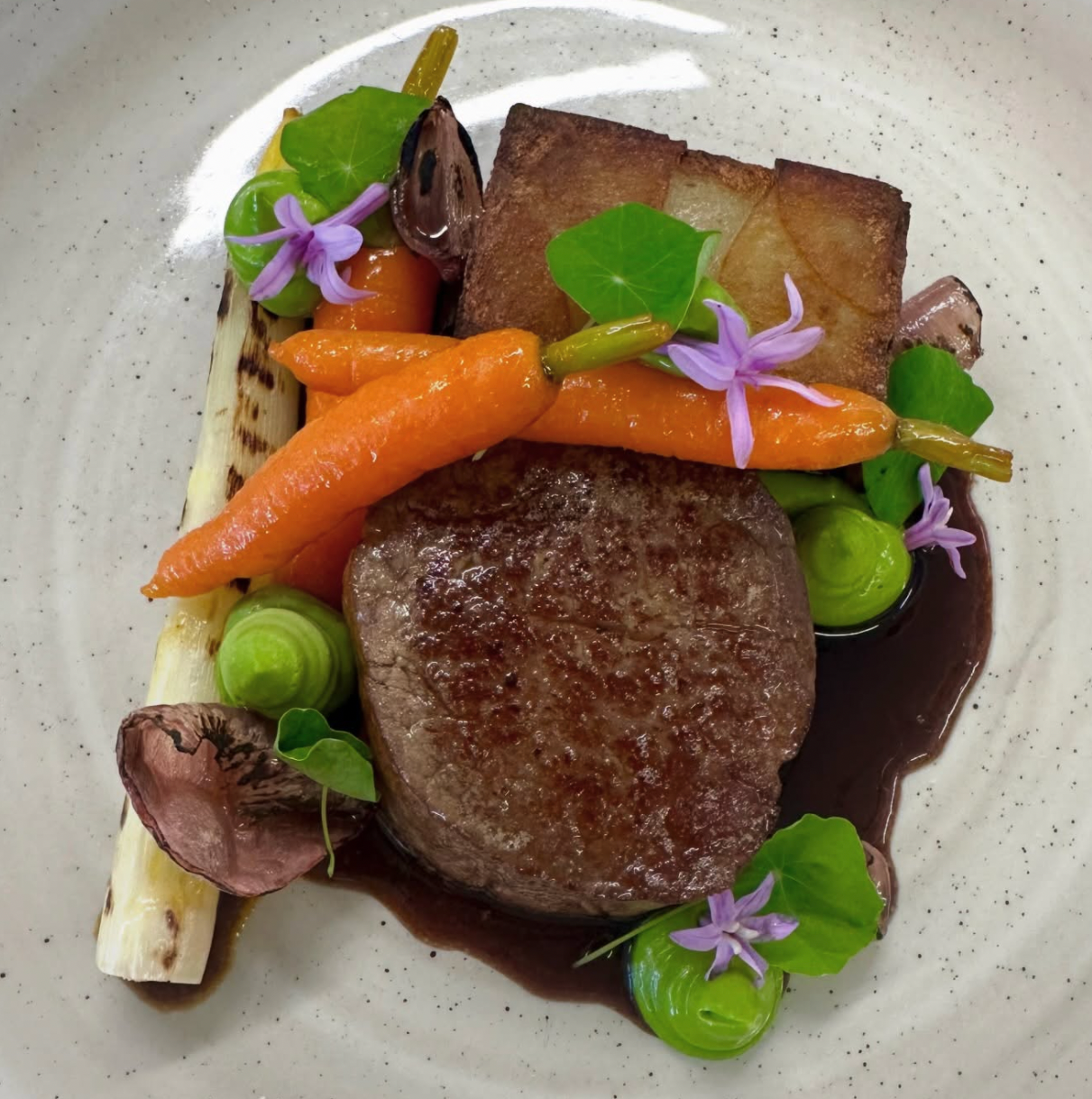 Seared steak with roasted carrots, charred abalone, grilled bread, microgreens, edible flowers, and sauce on a white plate.