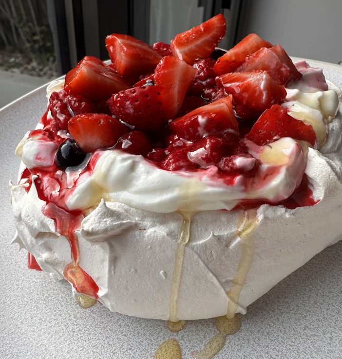 Pavlova featuring whipped cream, fresh strawberries, and mixed berries with drizzles of syrup on a white meringue base.