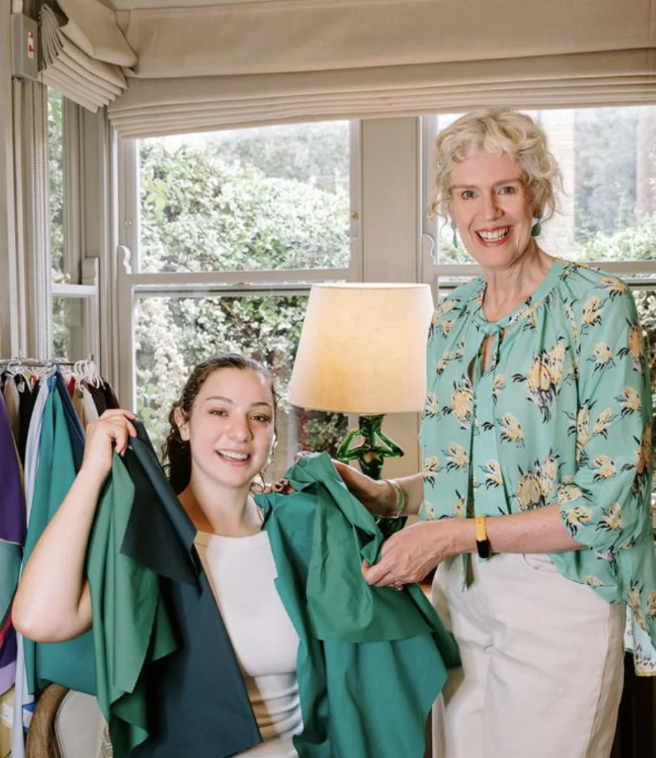 A woman with curly blonde hair handing a teal piece of clothing to a younger woman with brown hair, who is sitting and showing a smile. They are surrounded by racks of colorful clothing in a room with large windows and greenery outside.