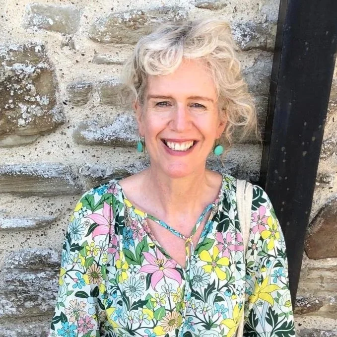 A smiling woman with short, curly hair, wearing teal earrings and a colorful floral blouse standing in front of a stone wall.