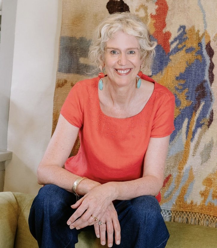 Woman with curly hair wearing a red top and turquoise earrings, sitting on a yellow couch with an abstract colorful tapestry behind her.